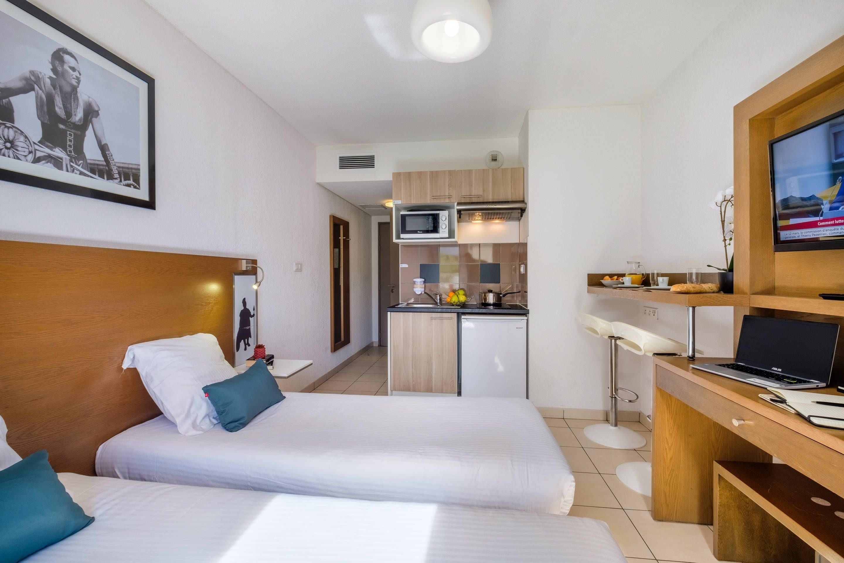 Een hotelkamer met twee bedden, een kitchenette, een televisie en een bureau met een laptop.