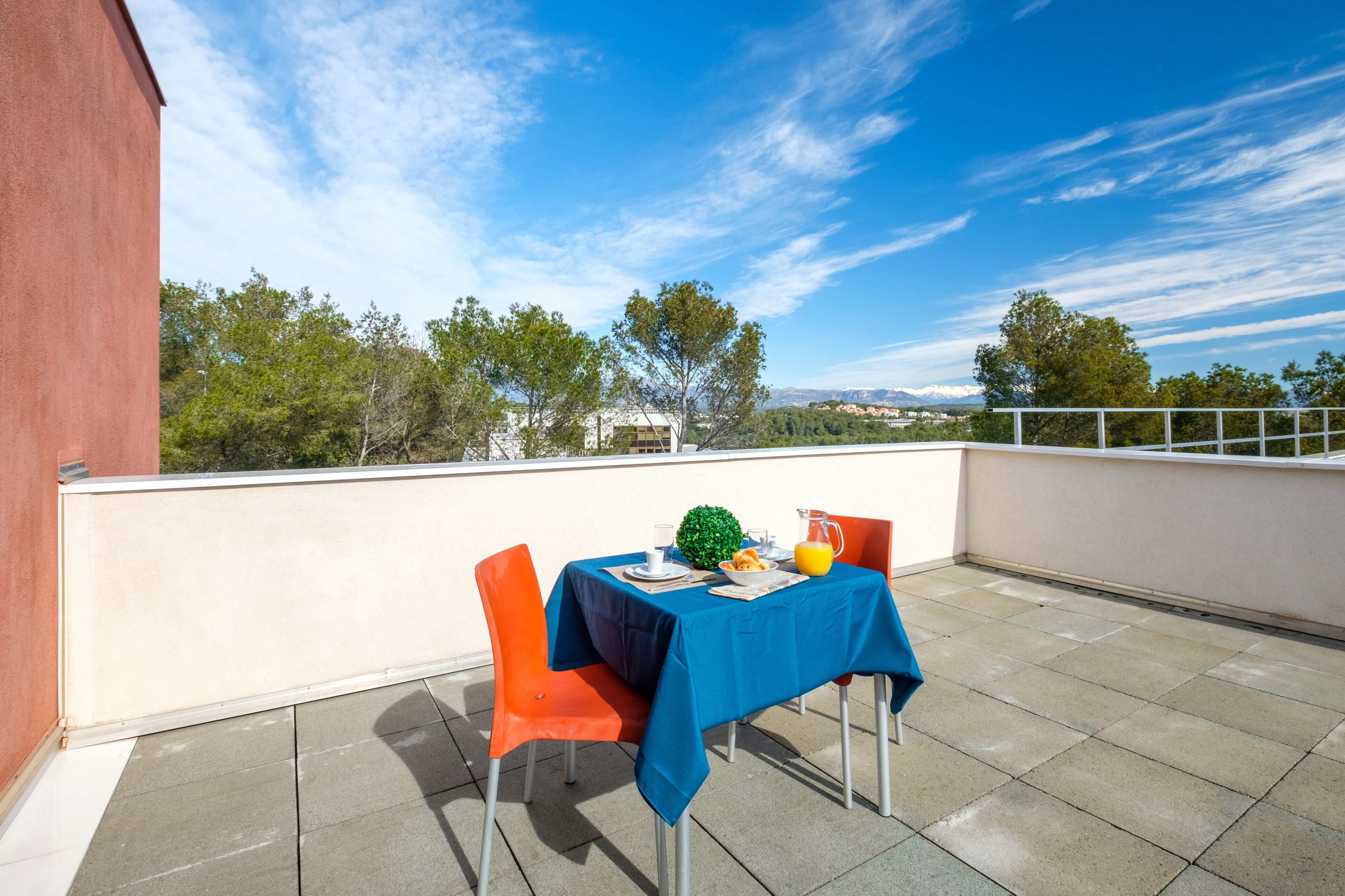 A terrace with a table set for two, a red building to the left, and a clear blue sky with trees and mountains in the background.