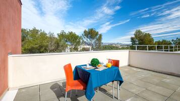 A terrace with a table set for two, a red building to the left, and a clear blue sky with trees and mountains in the background.