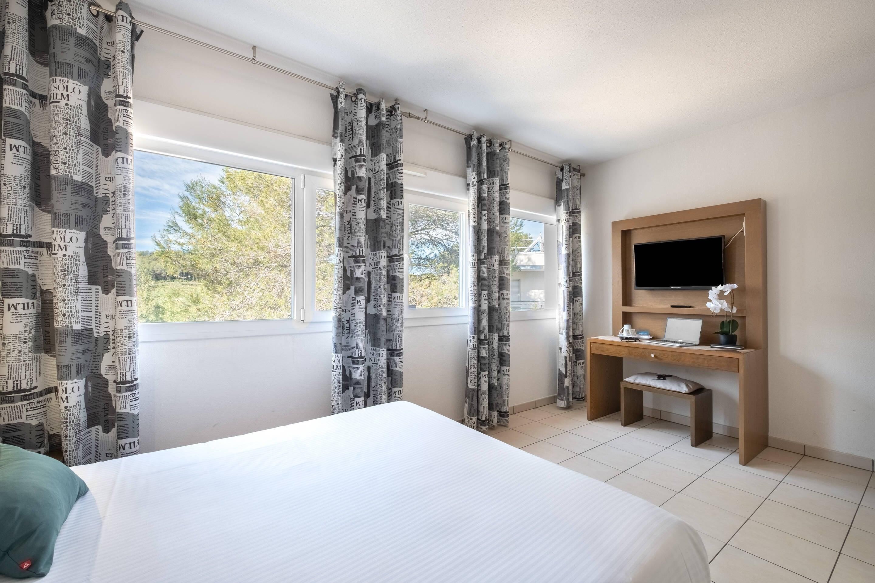 A hotel room with a bed, a desk with a computer, a TV, and large windows with curtains.