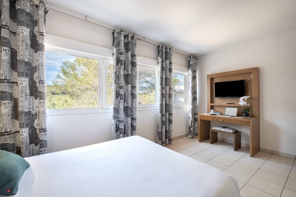 A hotel room with a bed, a desk with a computer, a TV, and large windows with curtains.