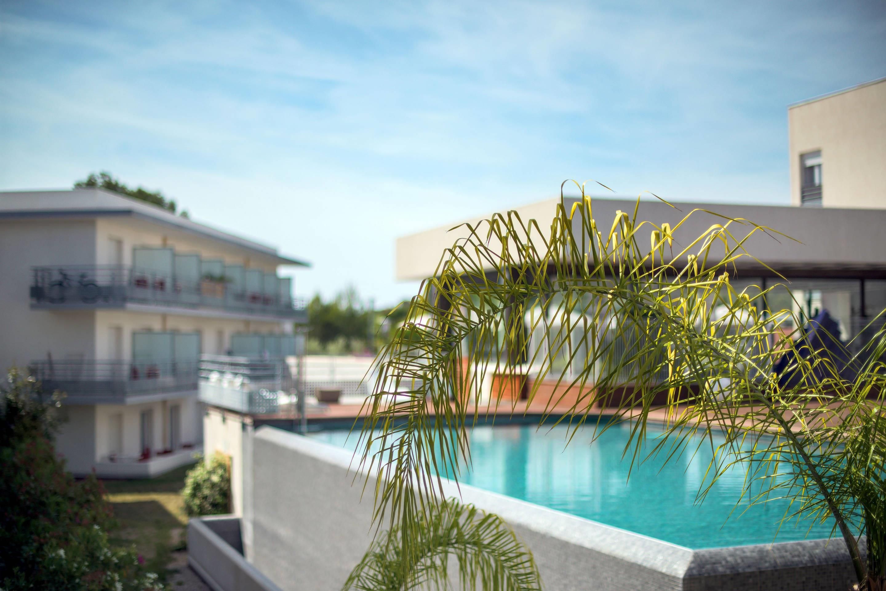 A swimming pool with a clear blue water surface, surrounded by a low stone wall. In the background, there are modern buildings with balconies.