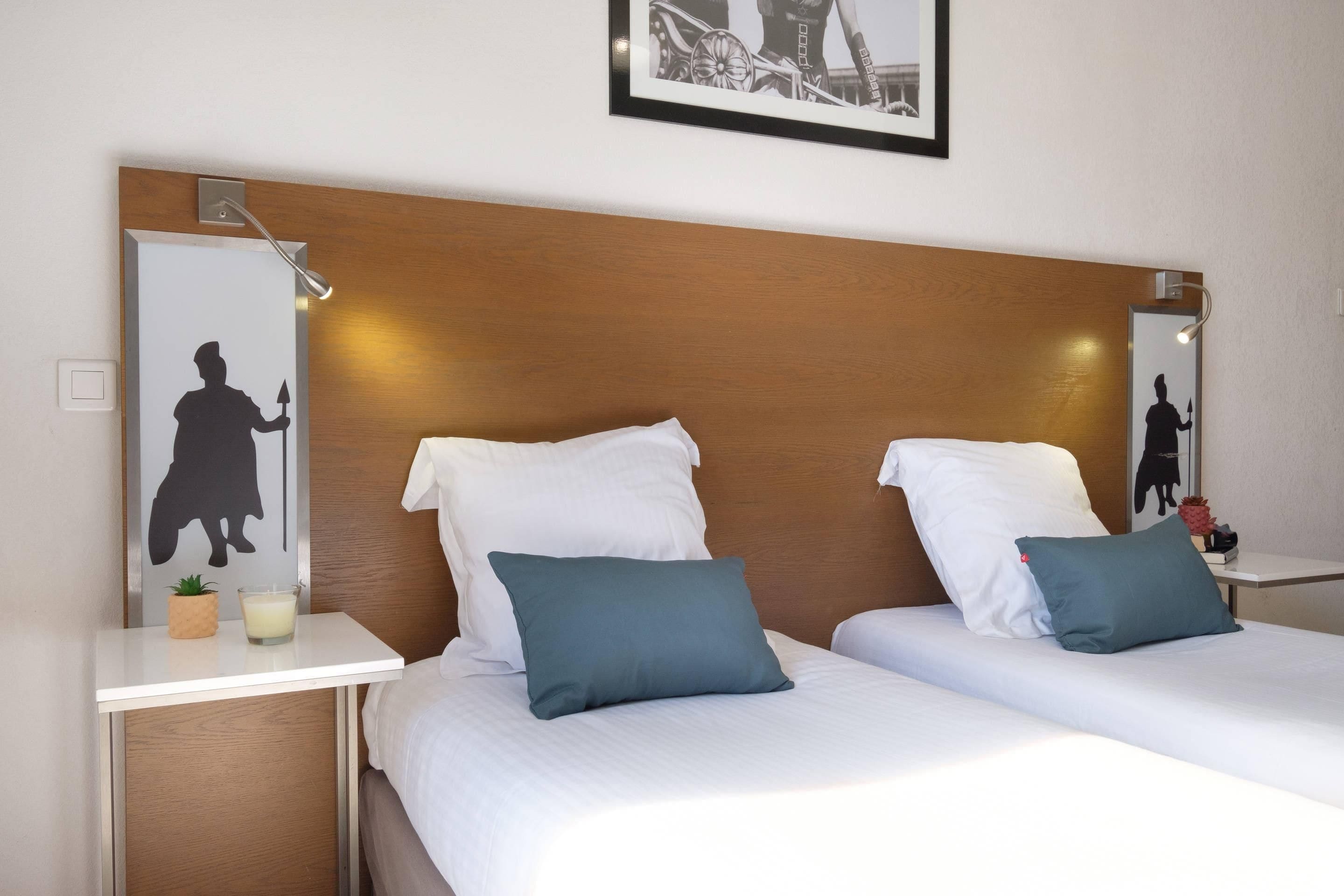 A hotel room with two beds, white bedding, teal pillows, a wooden headboard, a bedside table with a lamp, a small plant, and a framed picture on the wall.