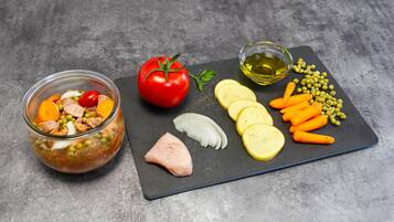A glass bowl of mixed ingredients, a whole tomato, sliced vegetables, and a small bowl of oil on a slate board.
