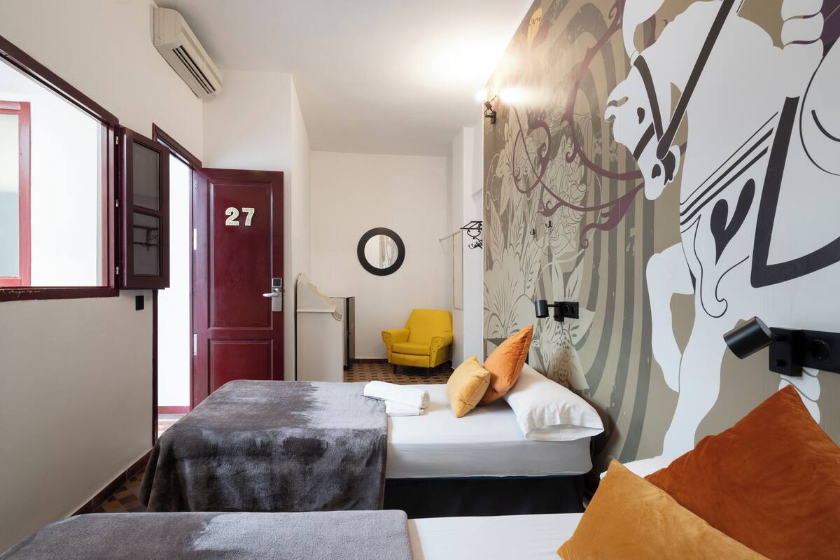 A hotel room with two beds, a yellow chair, a door labeled '27', and a wall with a horse mural.