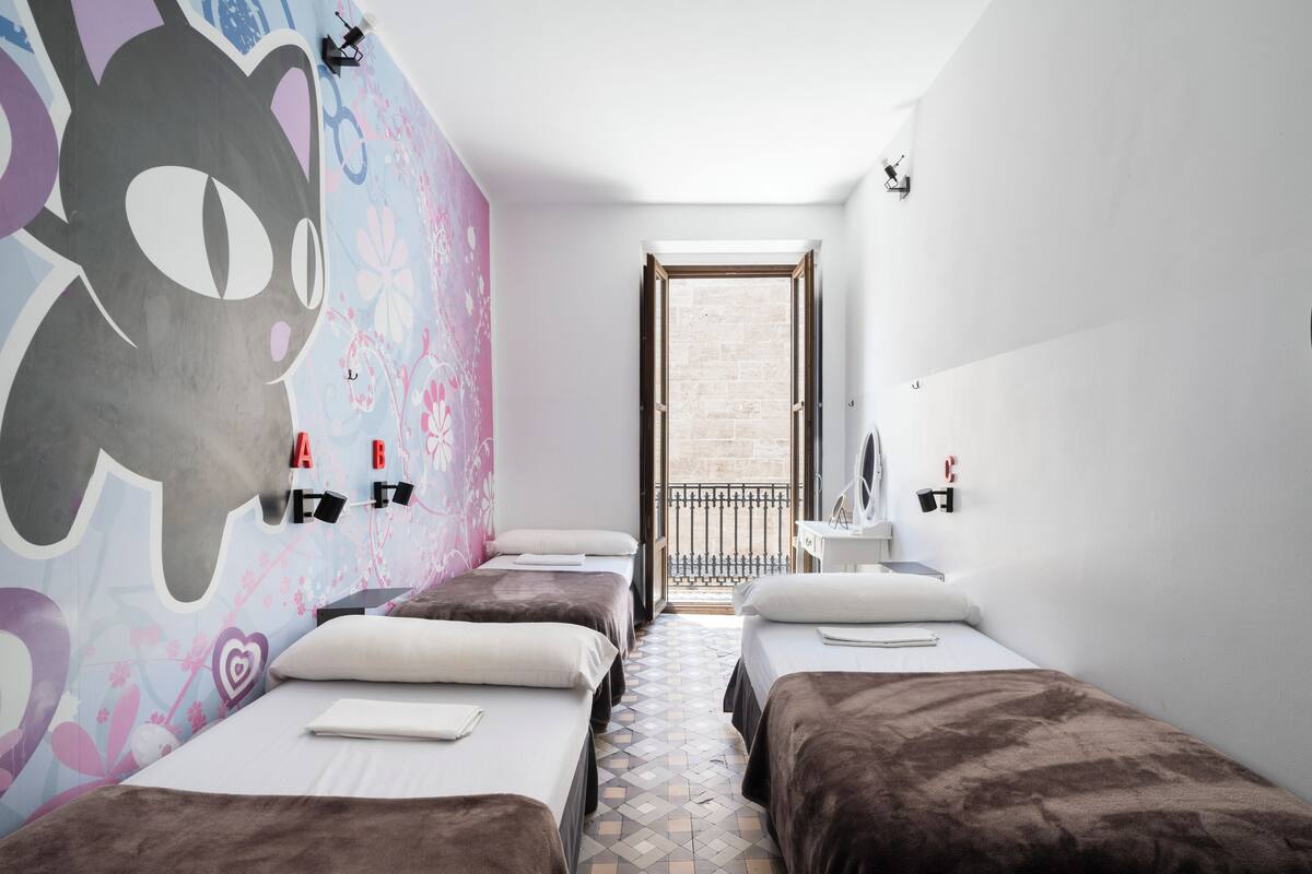 A hotel room with three beds, a mural on the left wall, a balcony with a view, and a small desk with a mirror and lamp.