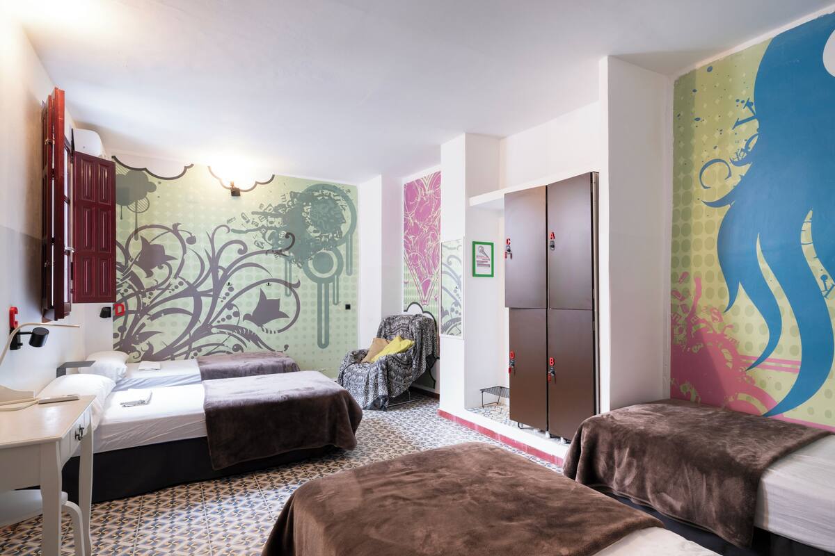 A room with two beds, a desk, a wardrobe, and a wall with a colorful mural.