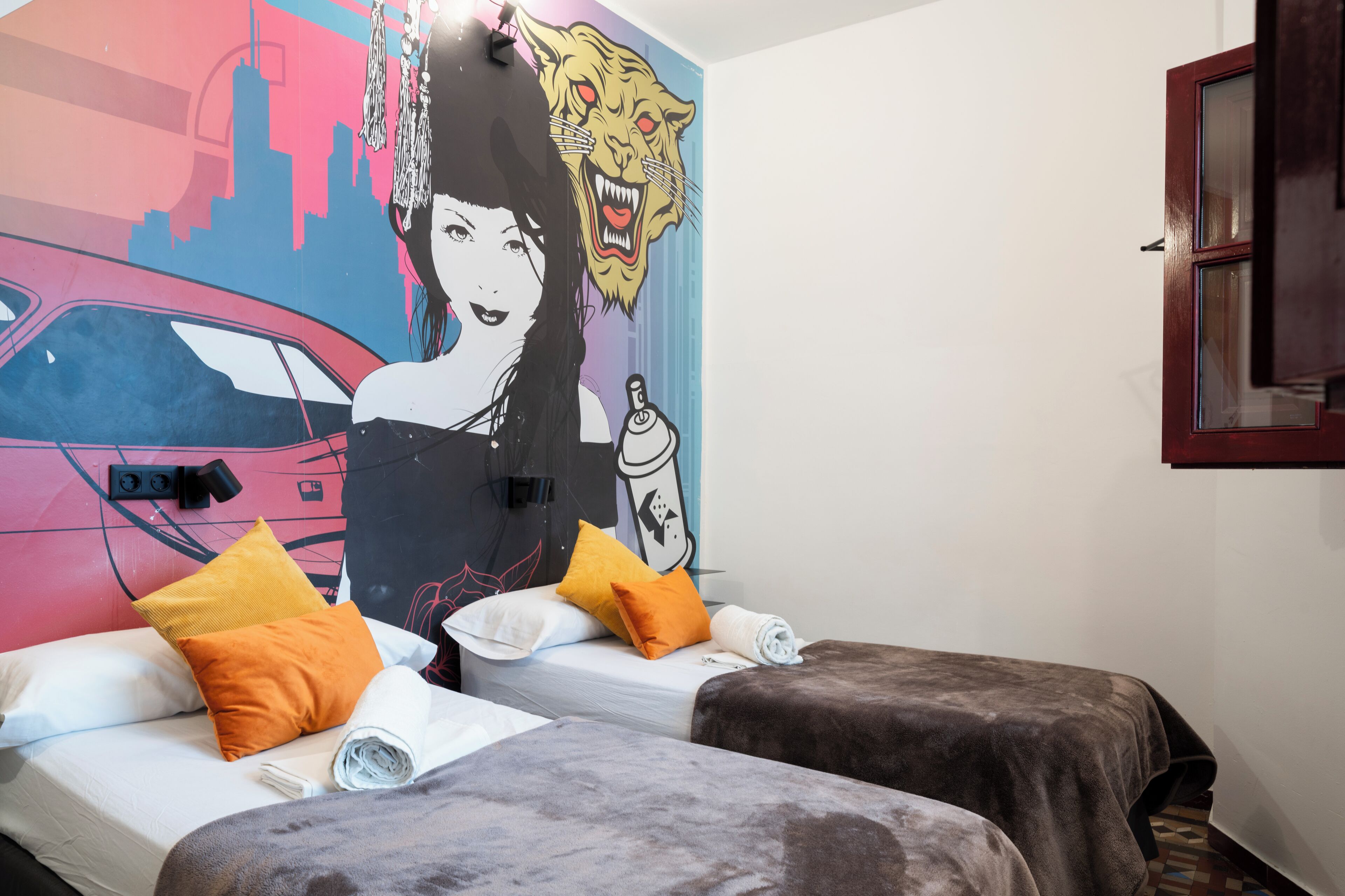 Ein Zimmer mit zwei Betten, jedes mit einer braunen Decke und orangefarbenen Kissen. Ein großes Wandbild zeigt eine Stadtansicht und einen Tiger.