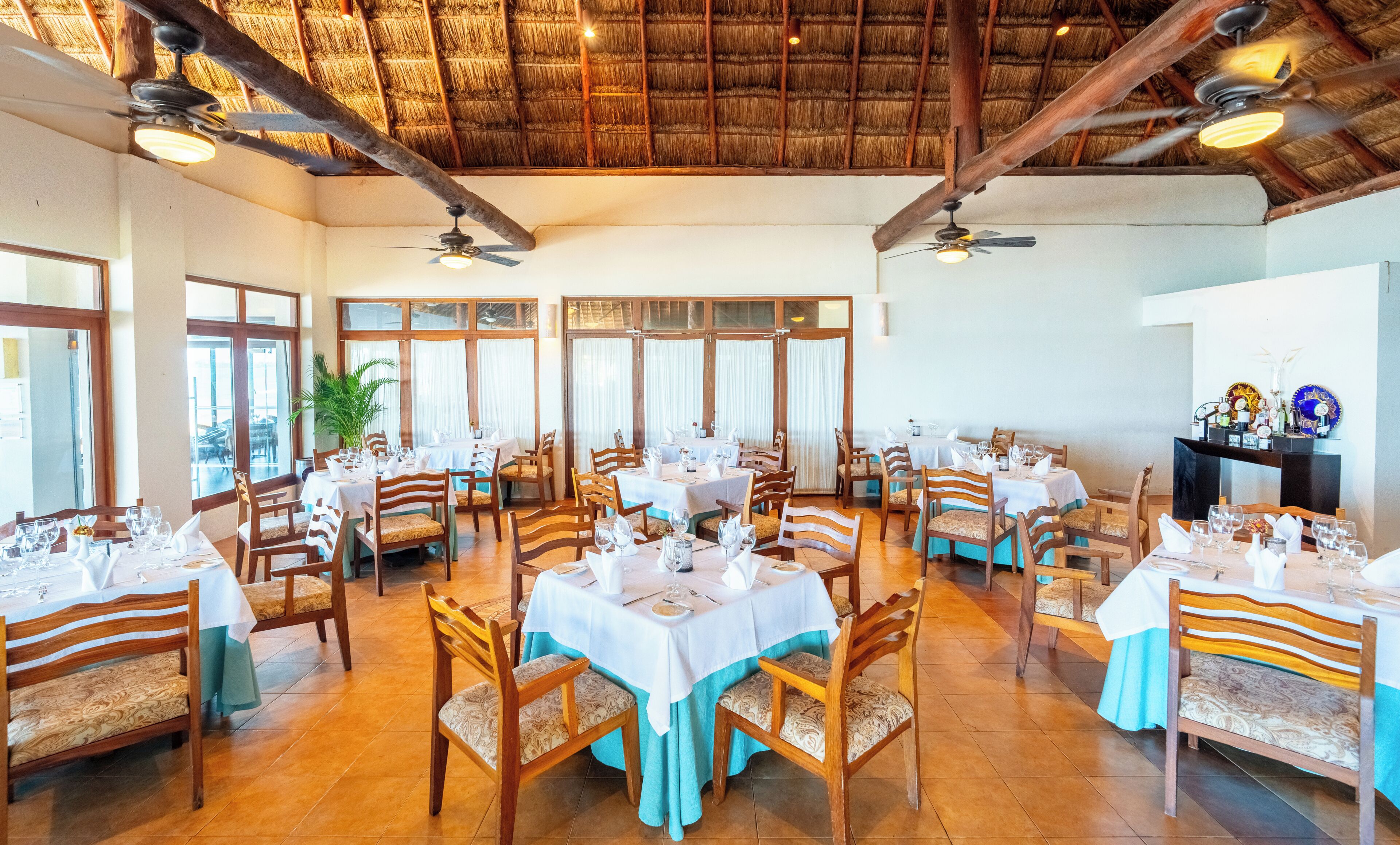 A restaurant with multiple tables, chairs, ceiling fans, and a thatched roof.