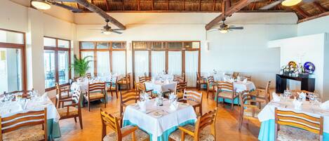 A restaurant with multiple tables, chairs, ceiling fans, and a thatched roof.