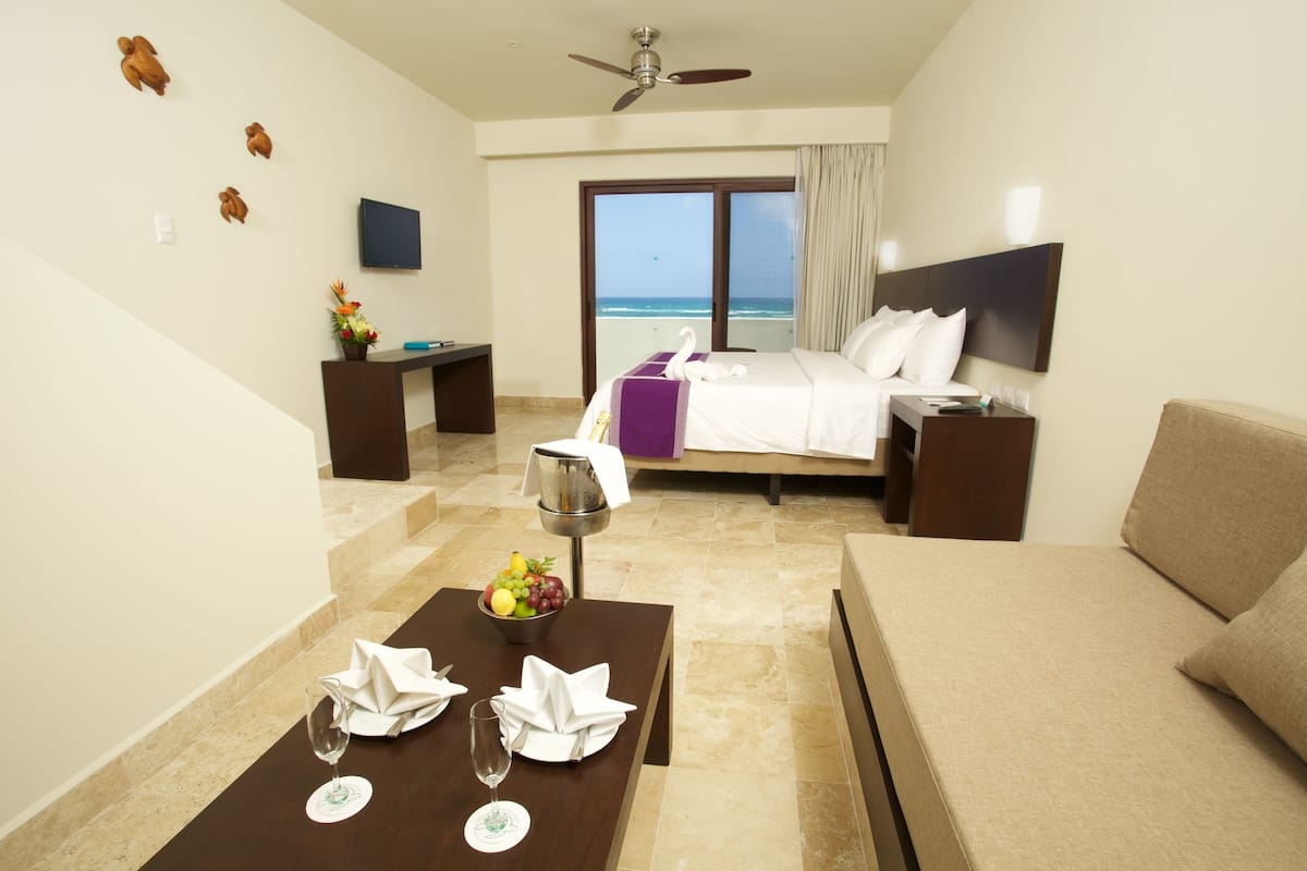Habitación de hotel con cama, escritorio, mesa de comedor y frutero, y vista al océano.