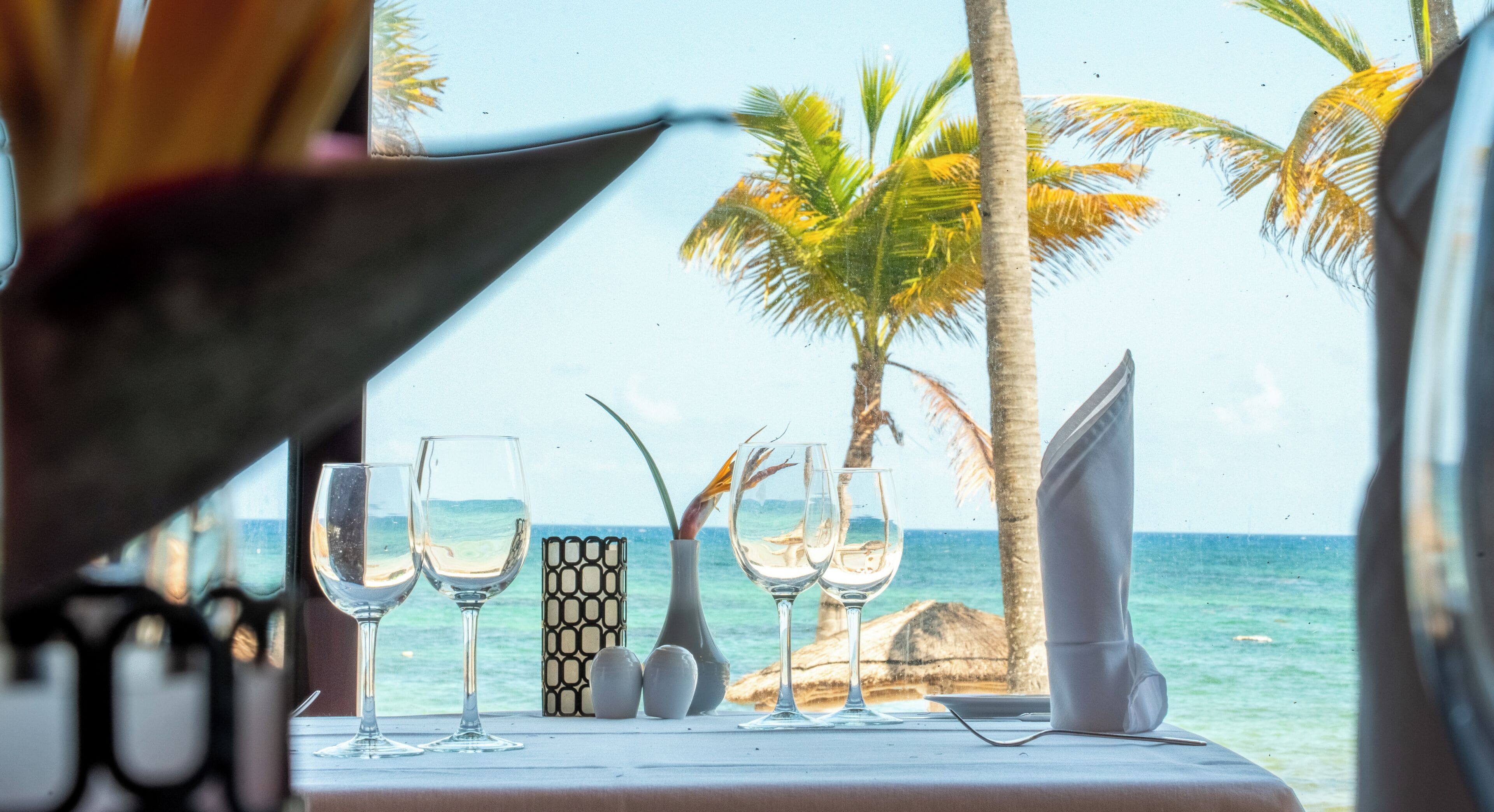 A table set for two with glasses and a vase, overlooking a beach with palm trees and the ocean.