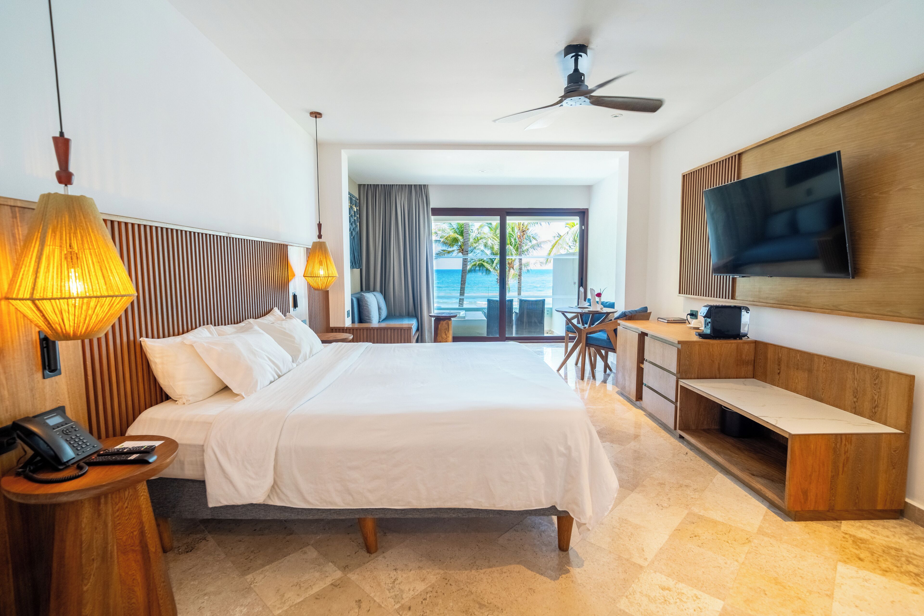 Habitación de hotel con cama, mesita de noche con teléfono, escritorio y silla, televisor montado en la pared, y vista al océano a través de la puerta de vidrio.
