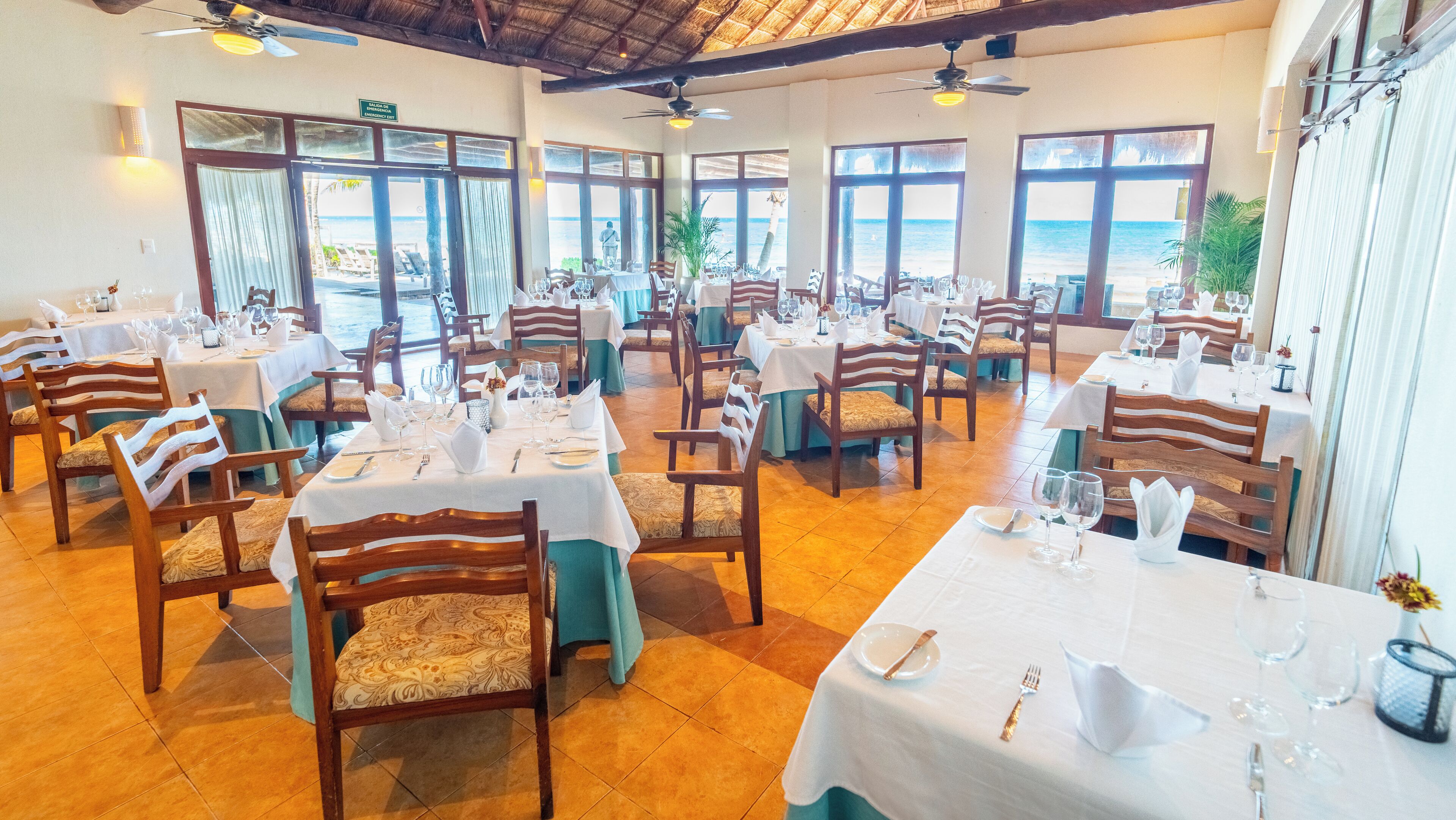 Un restaurant avec des tables dressées pour le service, des chaises en bois et de grandes fenêtres donnant sur l’océan.