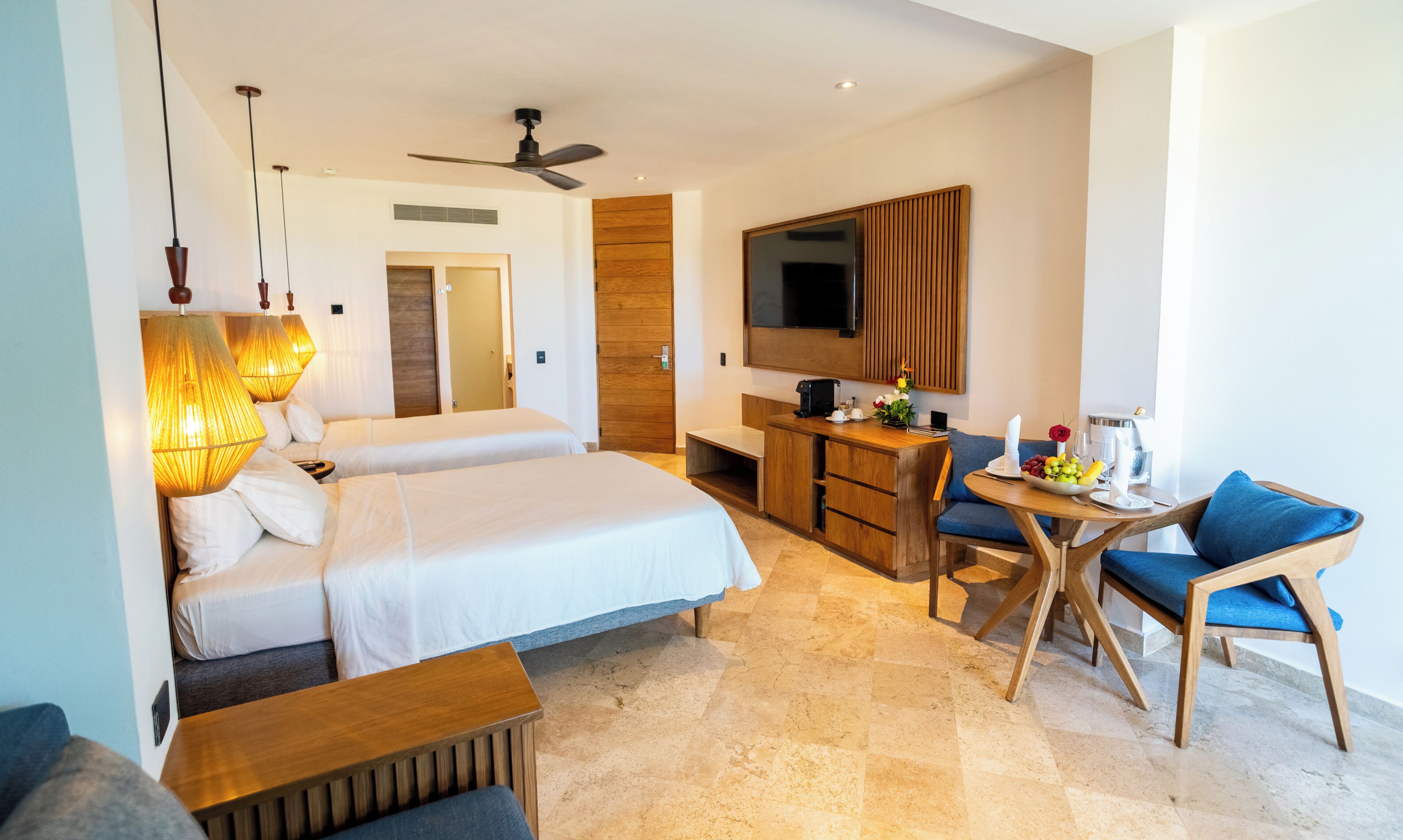 A hotel room with two beds, a desk, a chair, a TV, and a ceiling fan.