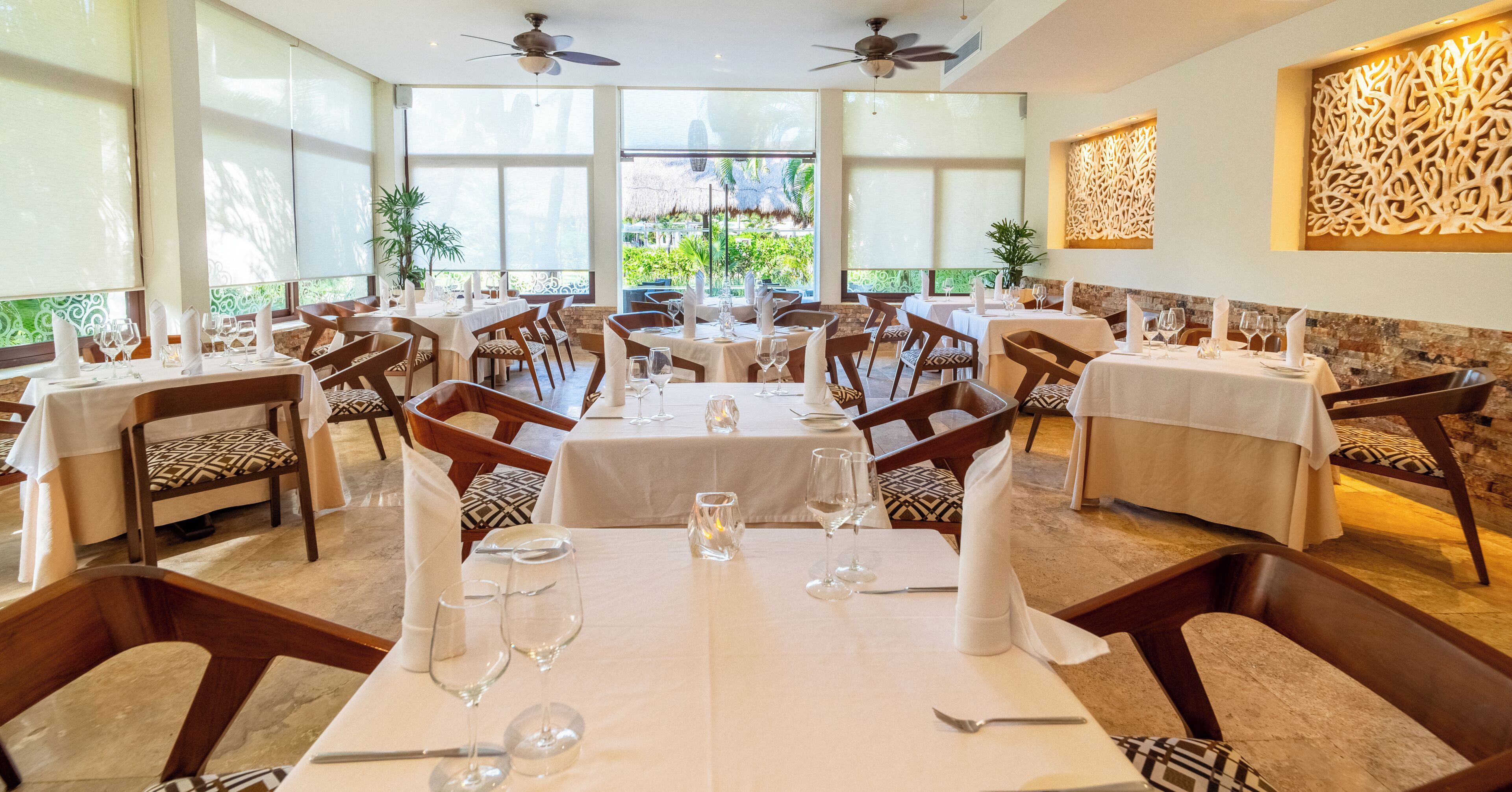 A restaurant with multiple tables, chairs, ceiling fans, and large windows with a view of greenery.