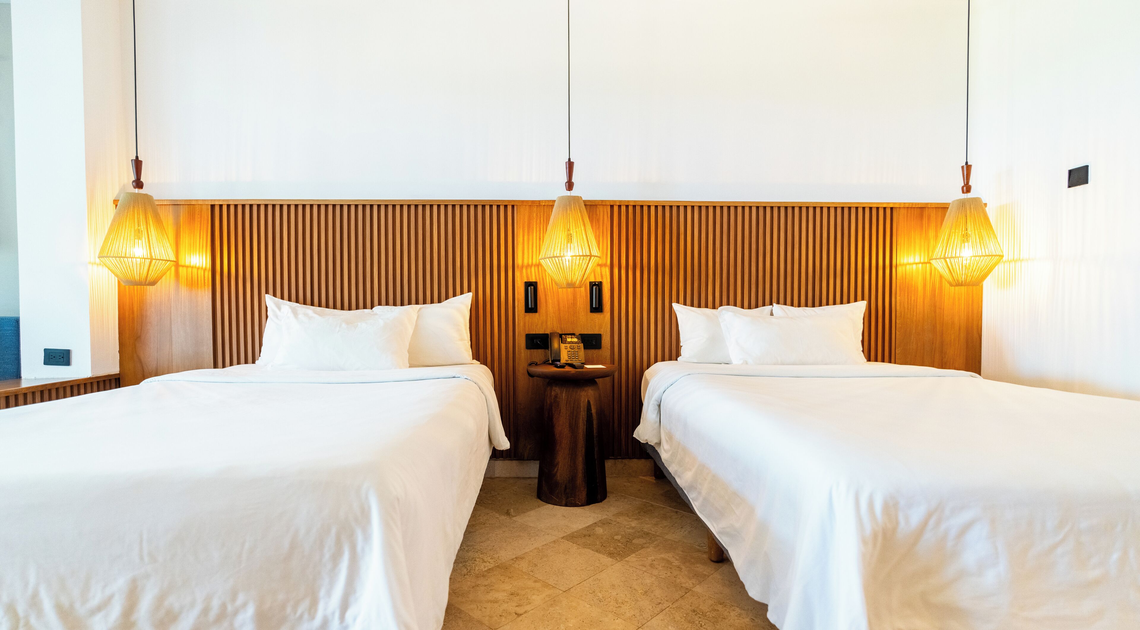 A hotel room with two beds, a wooden headboard, a bedside table with a phone, and wall-mounted lamps.