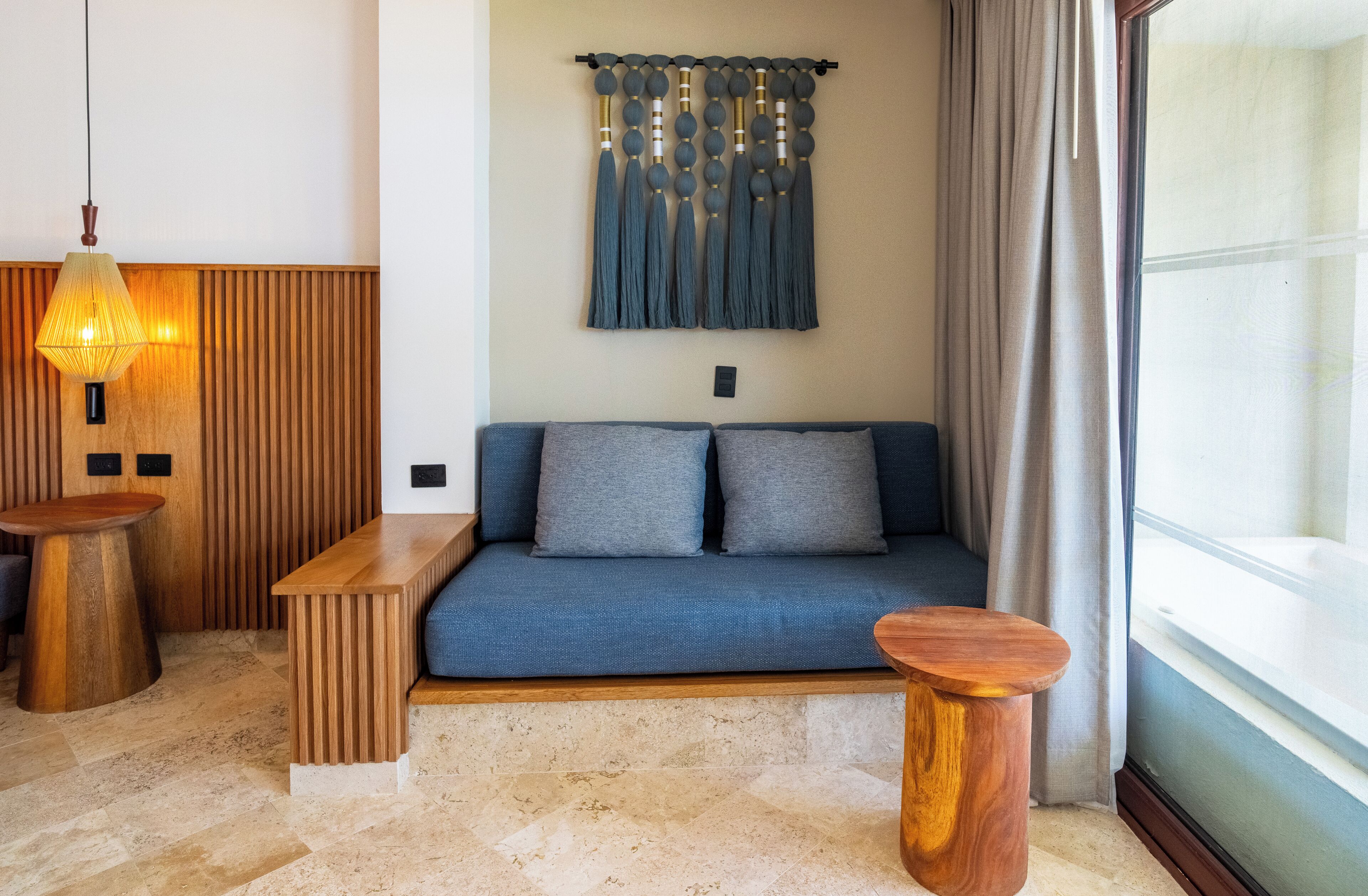 A modern living room with a blue sofa, wooden side tables, a hanging light fixture, and a wall-mounted curtain.