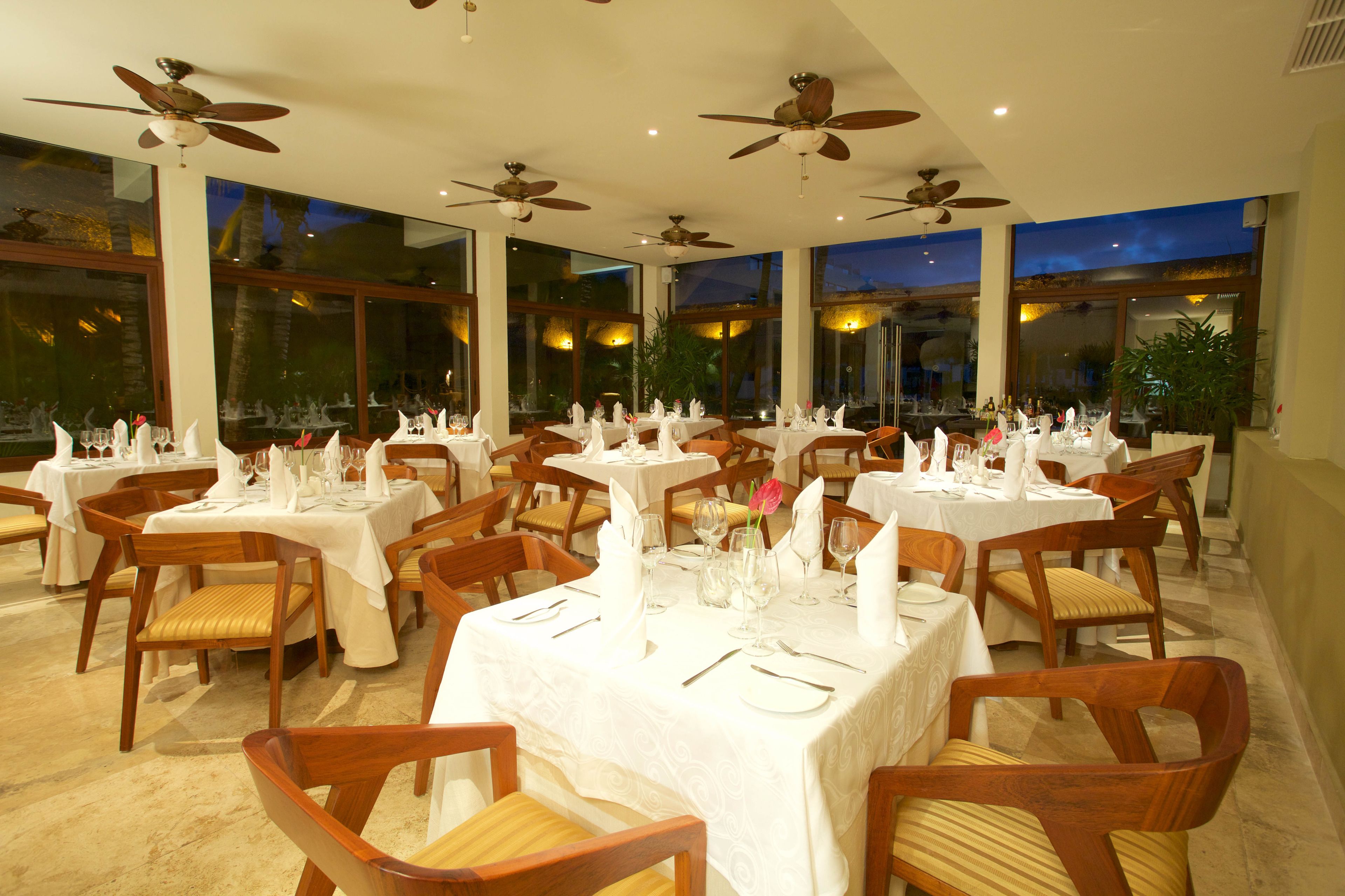 Un restaurante con varias mesas dispuestas para comer, ventiladores de techo y grandes ventanales.