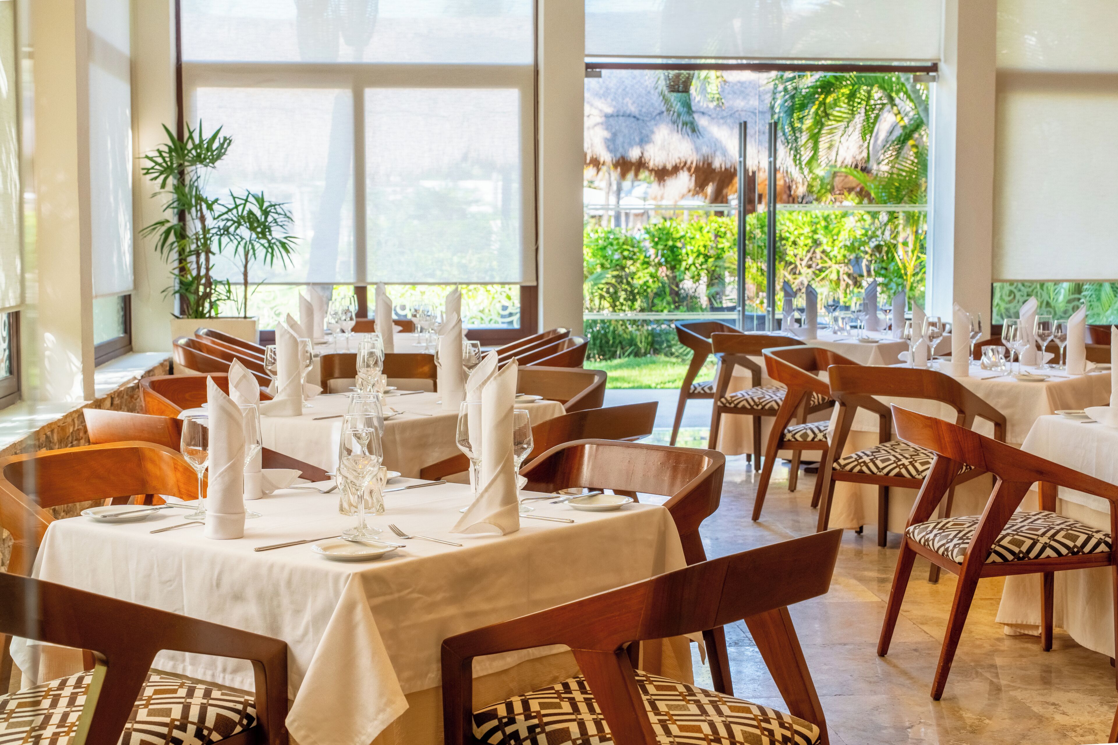 Un restaurant avec des tables dressées, des chaises et une grande fenêtre donnant sur un paysage verdoyant.