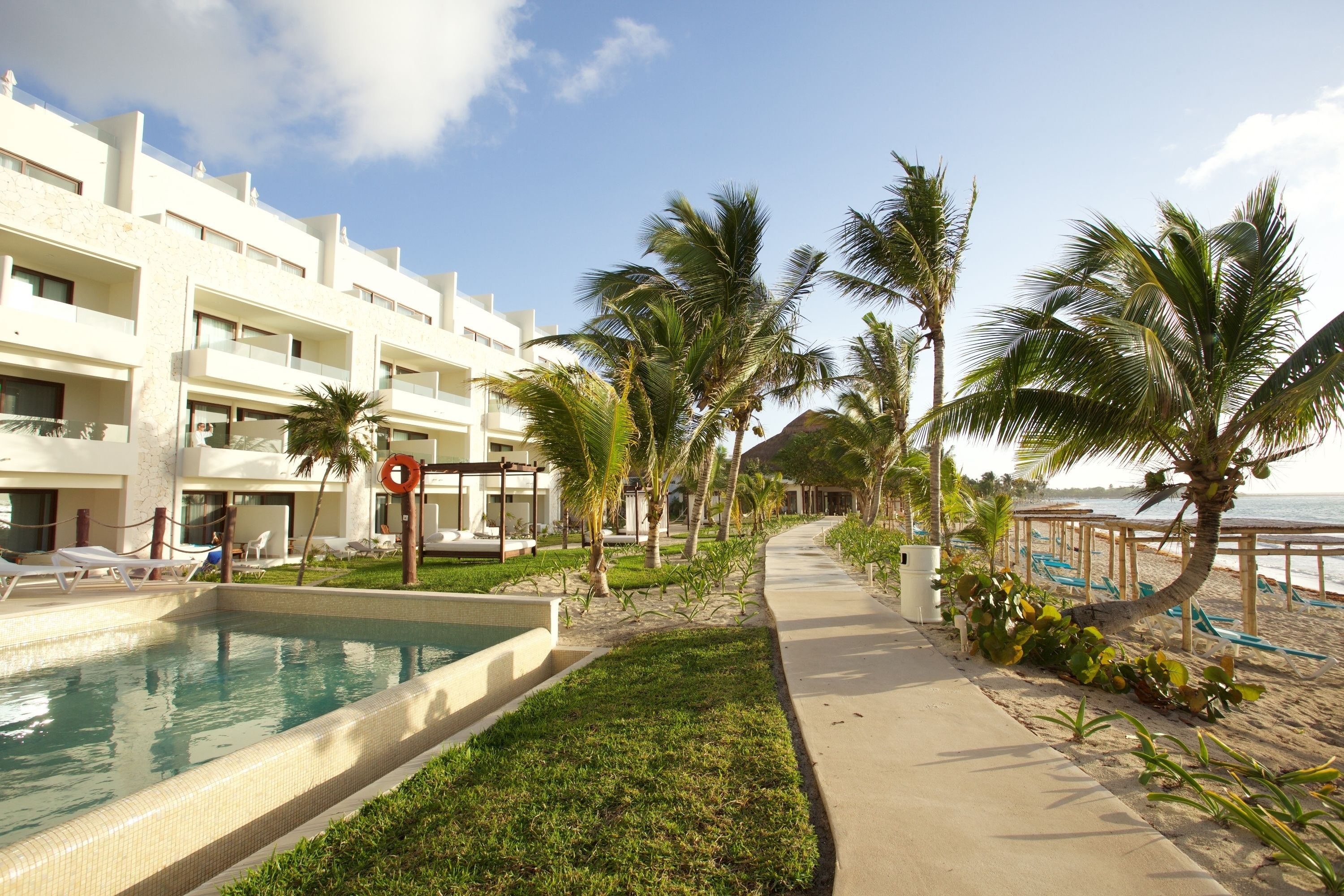 A pool area with a clear view of the sea, a pathway lined with palm trees, and a building with multiple balconies.