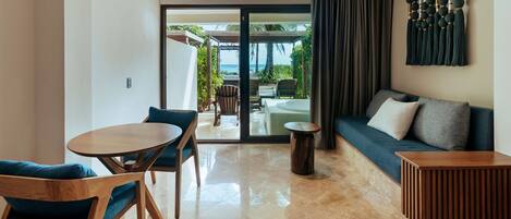 Une chambre d’hôtel moderne avec une table et des chaises en bois, un canapé avec des coussins, une petite table d’appoint en bois et une vue sur l’océan par une porte vitrée.