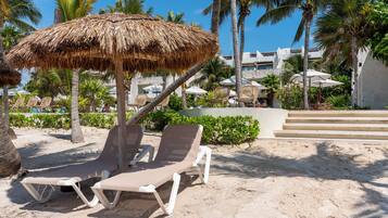 Un resort frente a la playa con sillas de descanso, una sombrilla de paja, palmeras y un edificio con escalones.