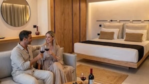 Una habitación de hotel moderna con un sofá, una mesa de centro redonda con fruta y vino, una cama con almohadas y un armario de madera.