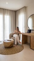 Une femme est assise sur une chaise dans une pièce où se trouvent une table ronde en bois, un ordinateur portable posé sur un bureau en bois et un grand miroir.