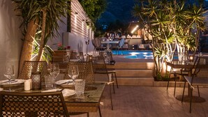Un área de comedor al aire libre con mesas y sillas, una piscina y montañas al fondo.