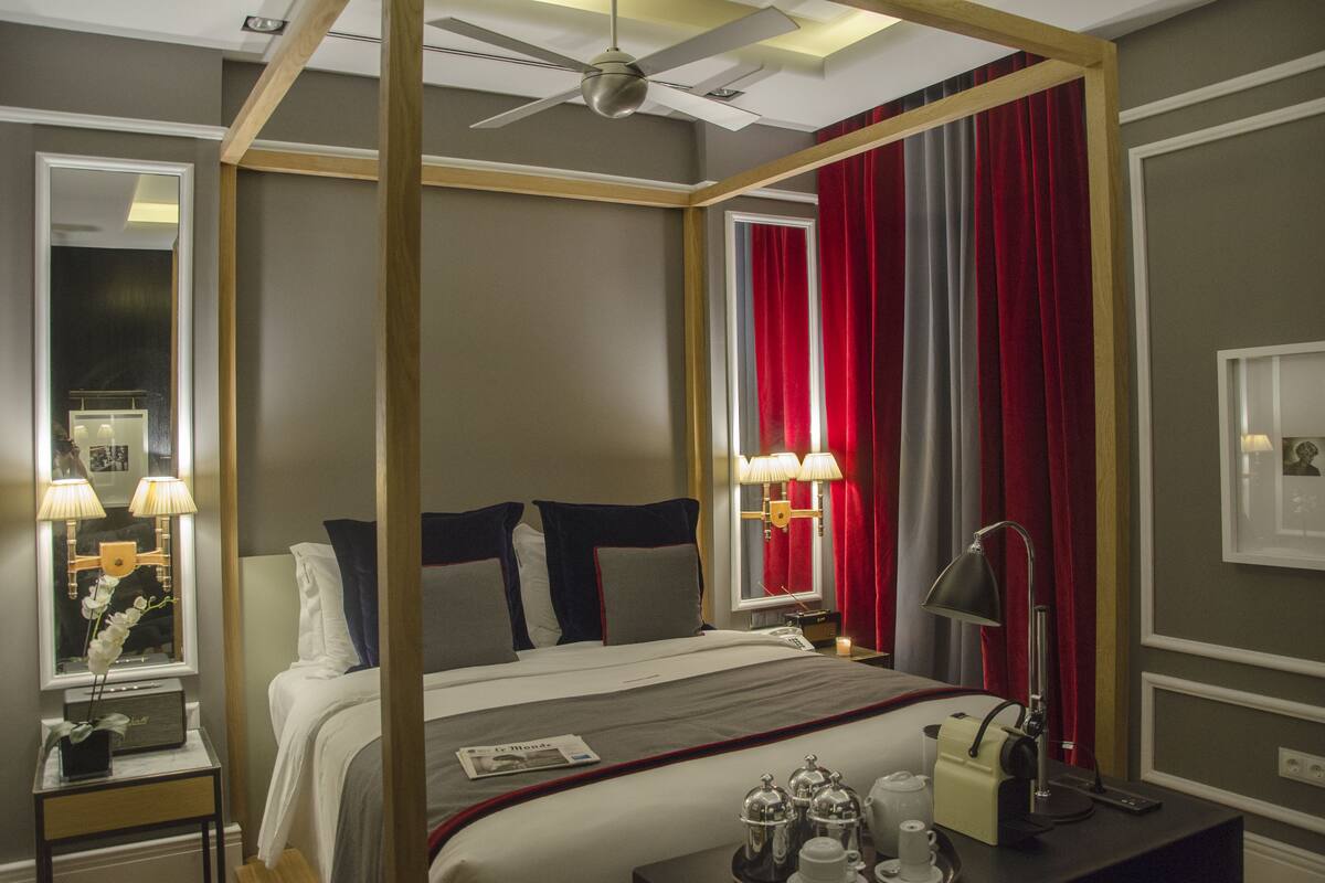 Una cama con dosel, con cubrecama gris y blanco, cortinas rojas y un ventilador de techo.
