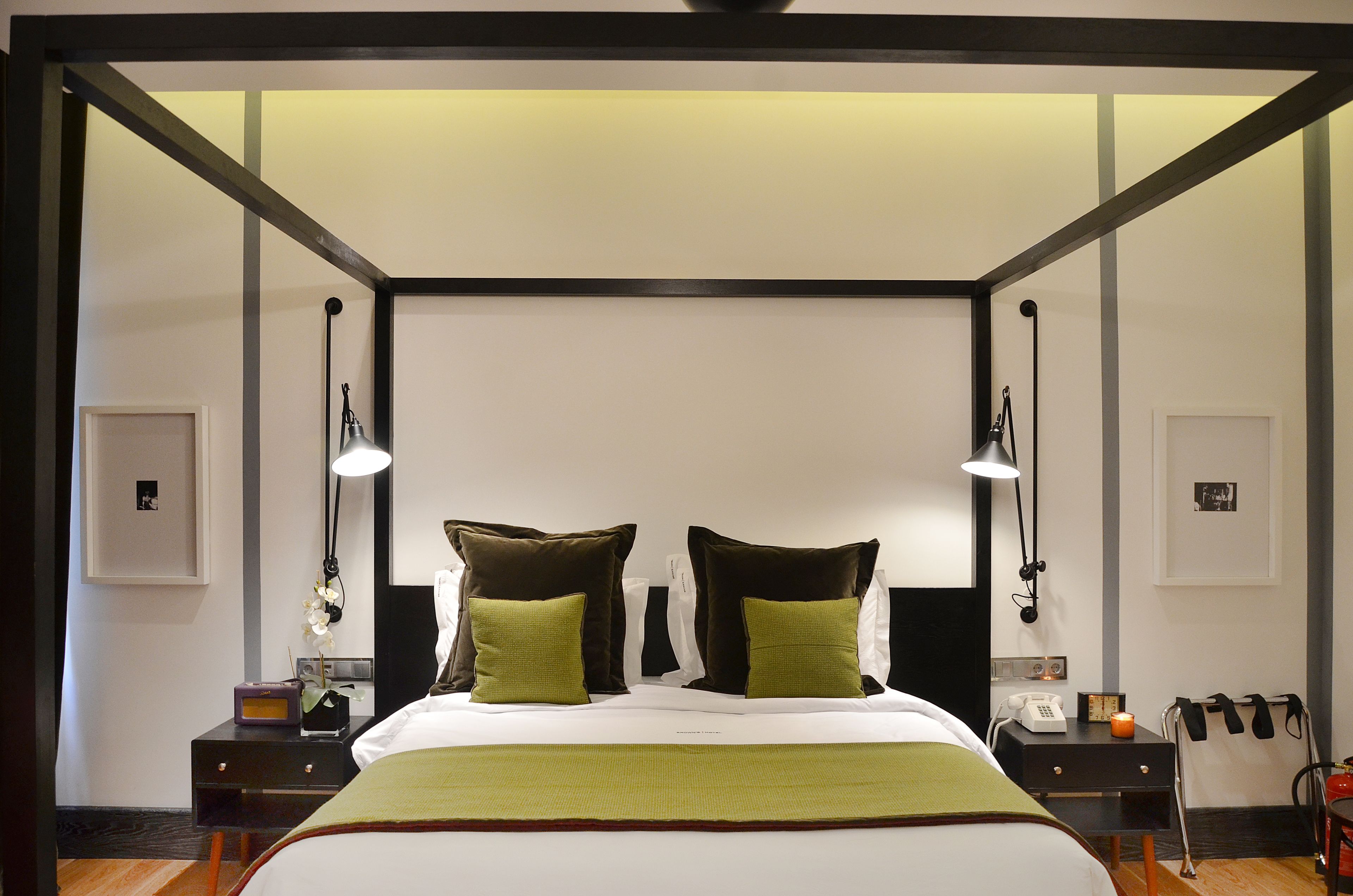 Une chambre d’hôtel avec un grand lit, deux lampes de chevet, une table de chevet avec une radio, un téléphone et une petite table avec une bougie.