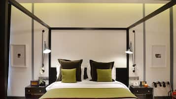 Une chambre d’hôtel avec un grand lit, deux lampes de chevet, une table de chevet avec une radio, un téléphone et une petite table avec une bougie.