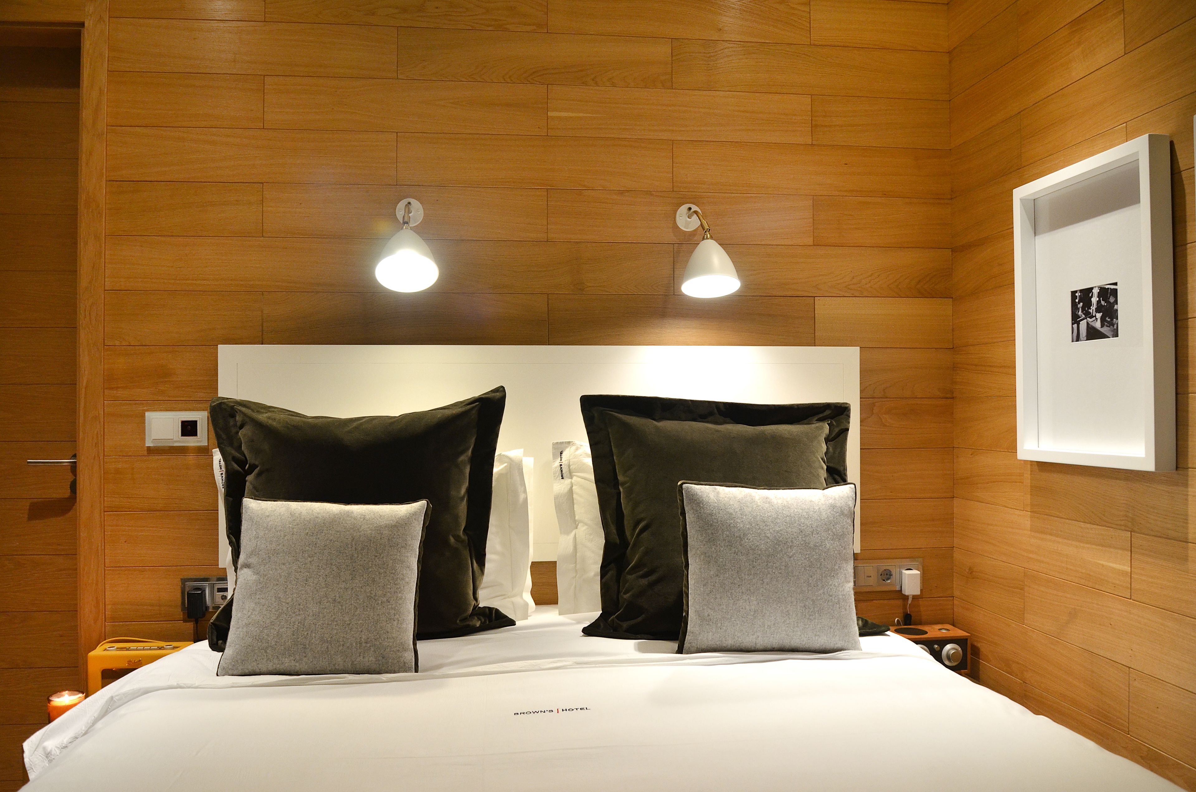 Quarto de hotel com teto de madeira, cama com roupa de cama branca, duas almofadas grandes, mesa de cabeceira com abajur e quadro emoldurado na parede.