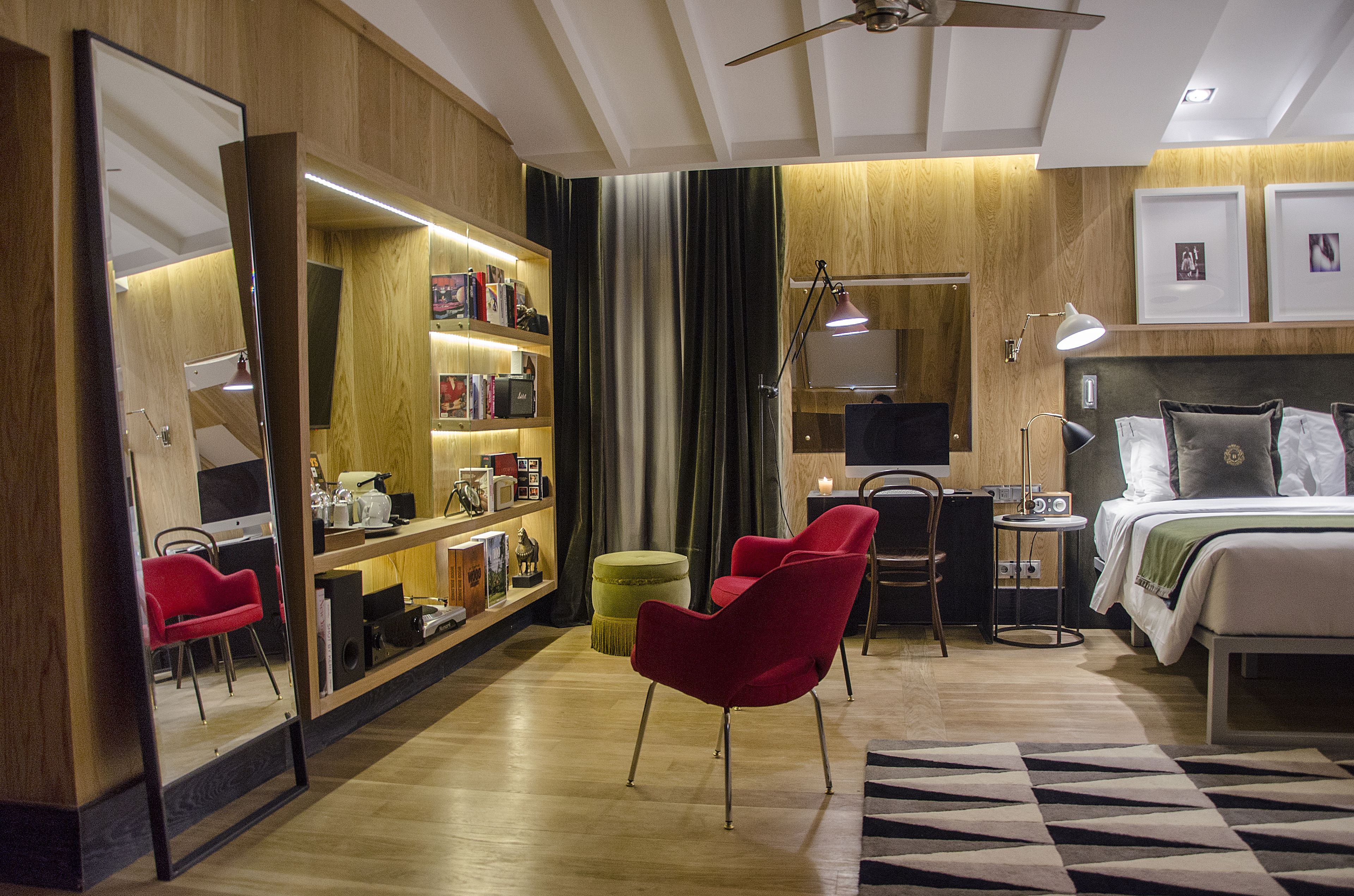 A modern hotel room with a wooden floor, a large bed with white bedding, a red armchair, a small round ottoman, a desk with a lamp, and a bookshelf filled with books and decorative items.