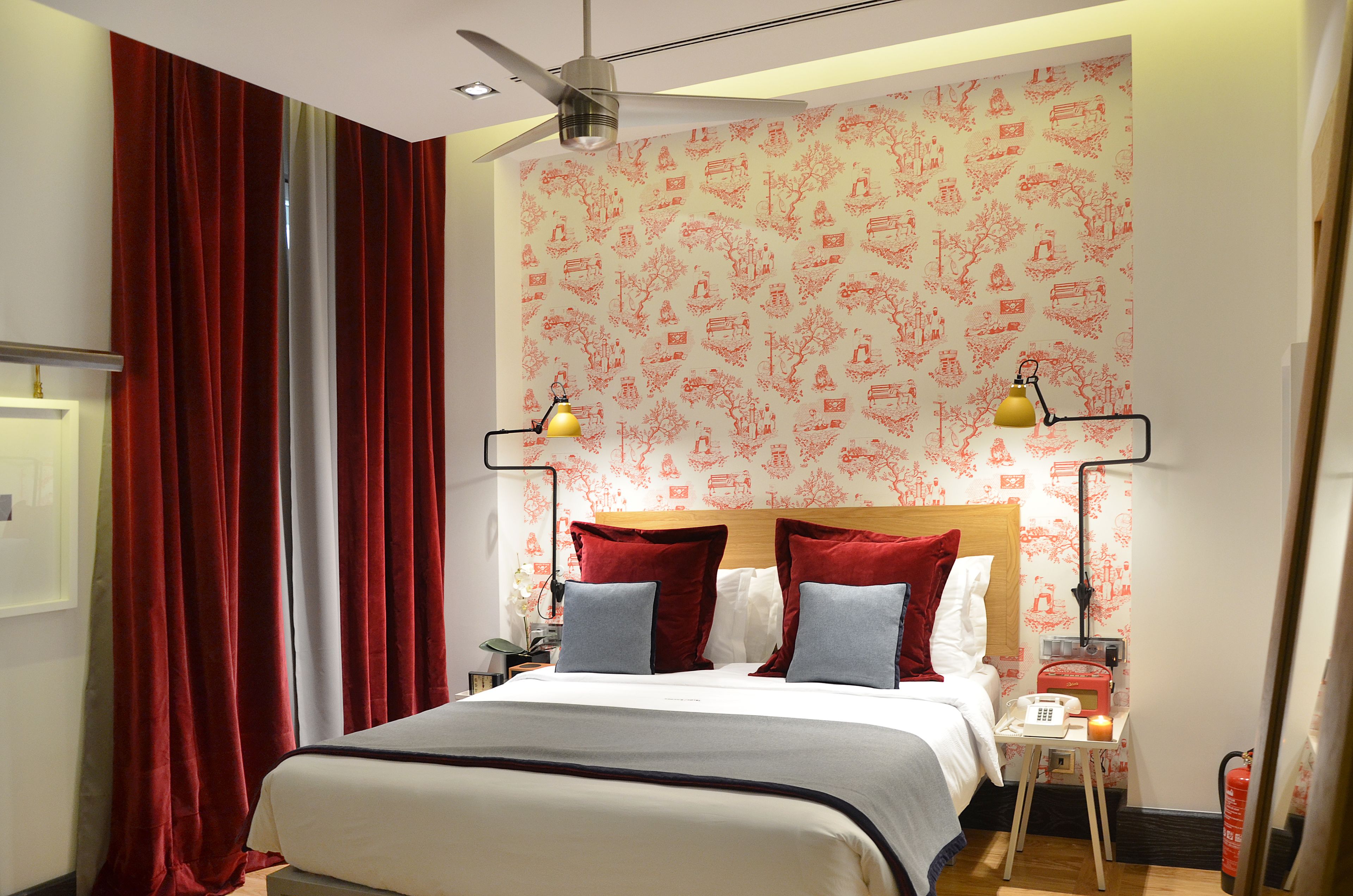 A hotel room with a bed, red curtains, a ceiling fan, a nightstand with a lamp, and a wall with red patterned wallpaper.