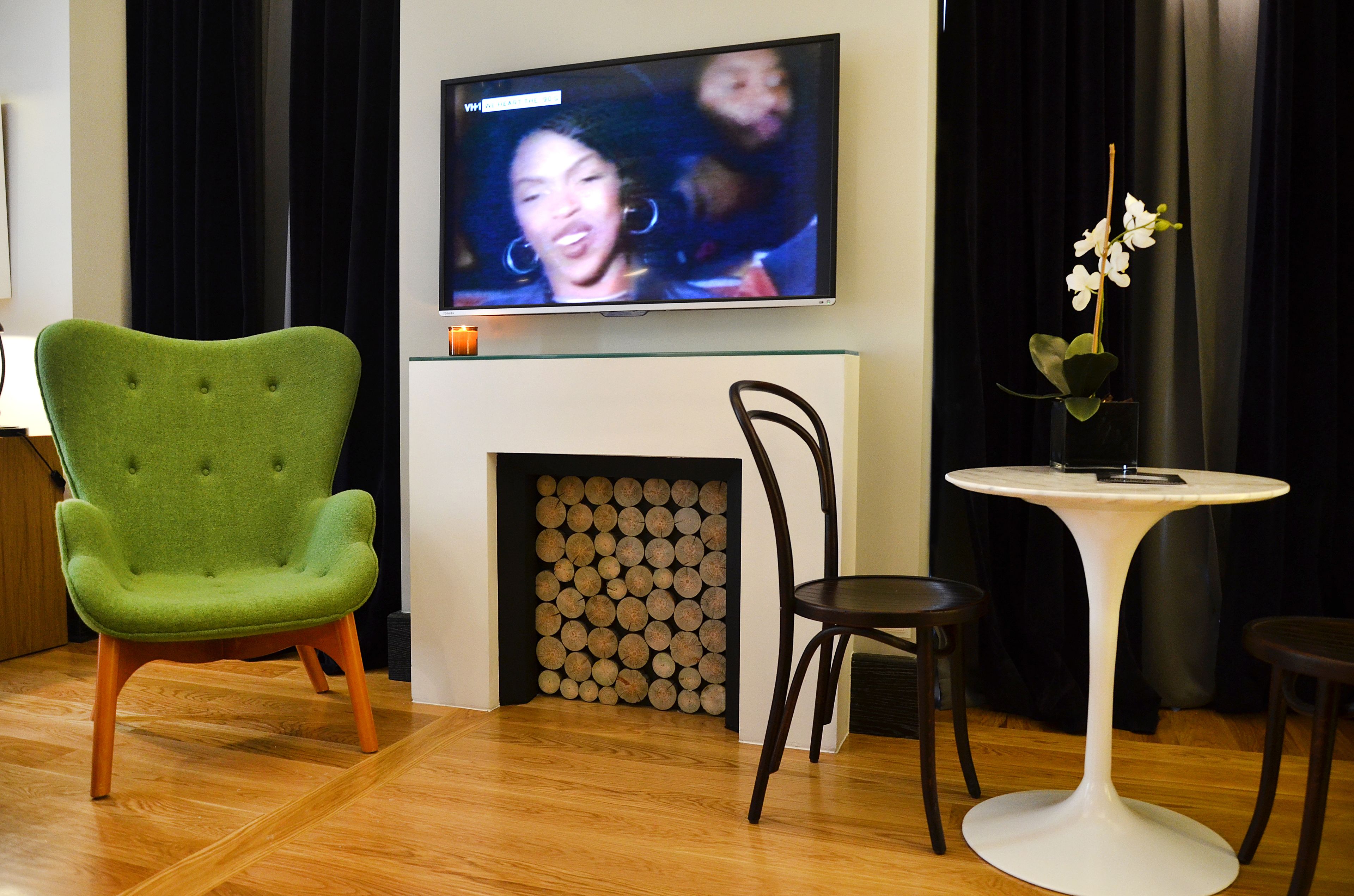 A living room with a green armchair, a black chair, a small round table, a fireplace with logs, and a TV mounted on the wall.