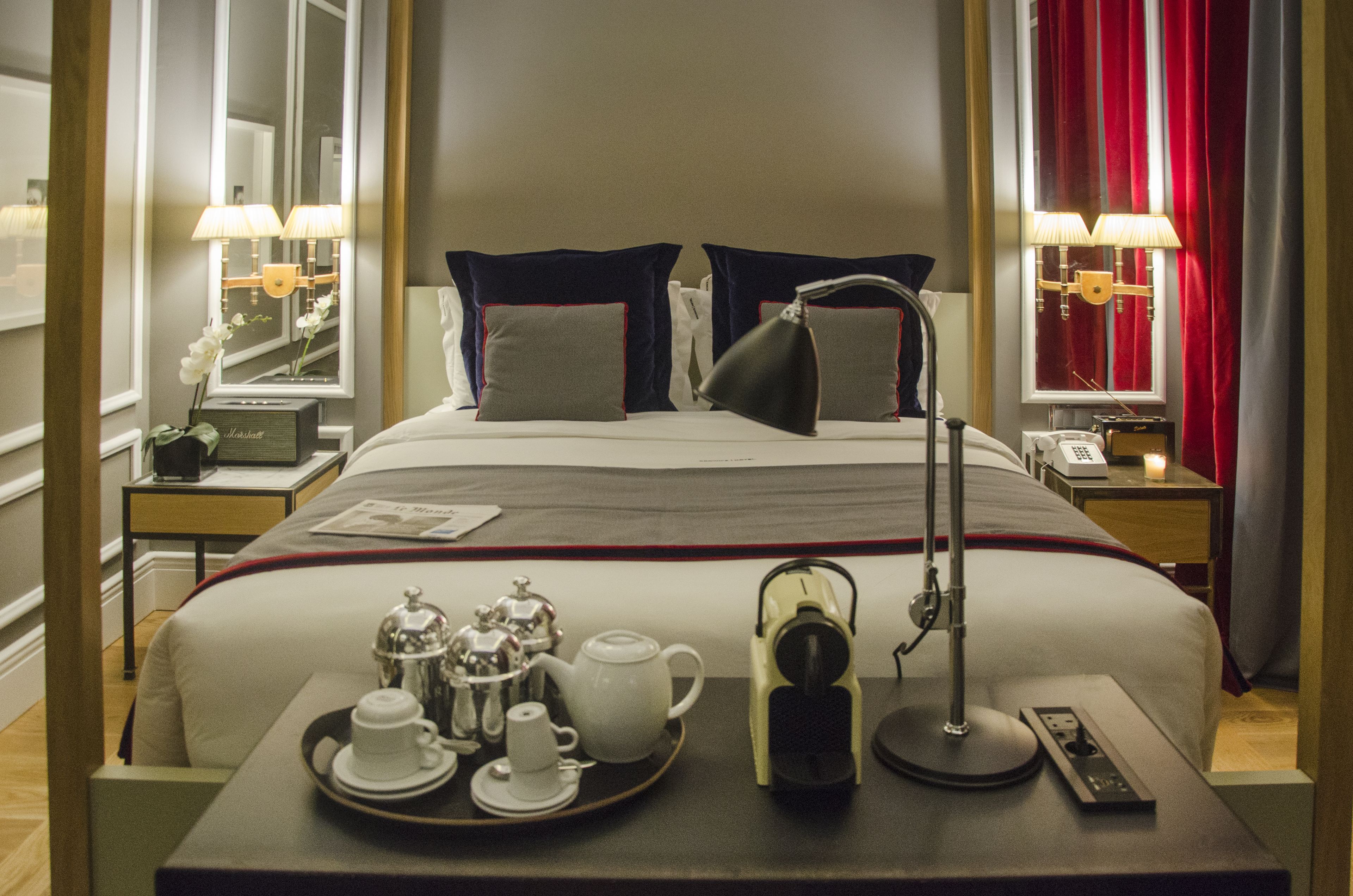 A hotel room with a neatly made bed, a tray with tea and cups, a bedside lamp, and a telephone.