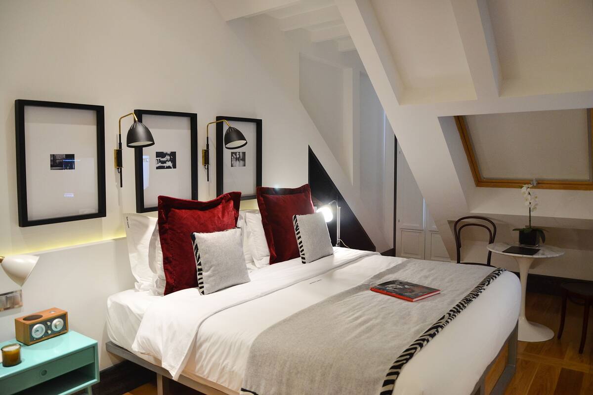 Un dormitorio ordenado con una cama, cuadros enmarcados en la pared, un escritorio con una lámpara y una mesita con una planta.