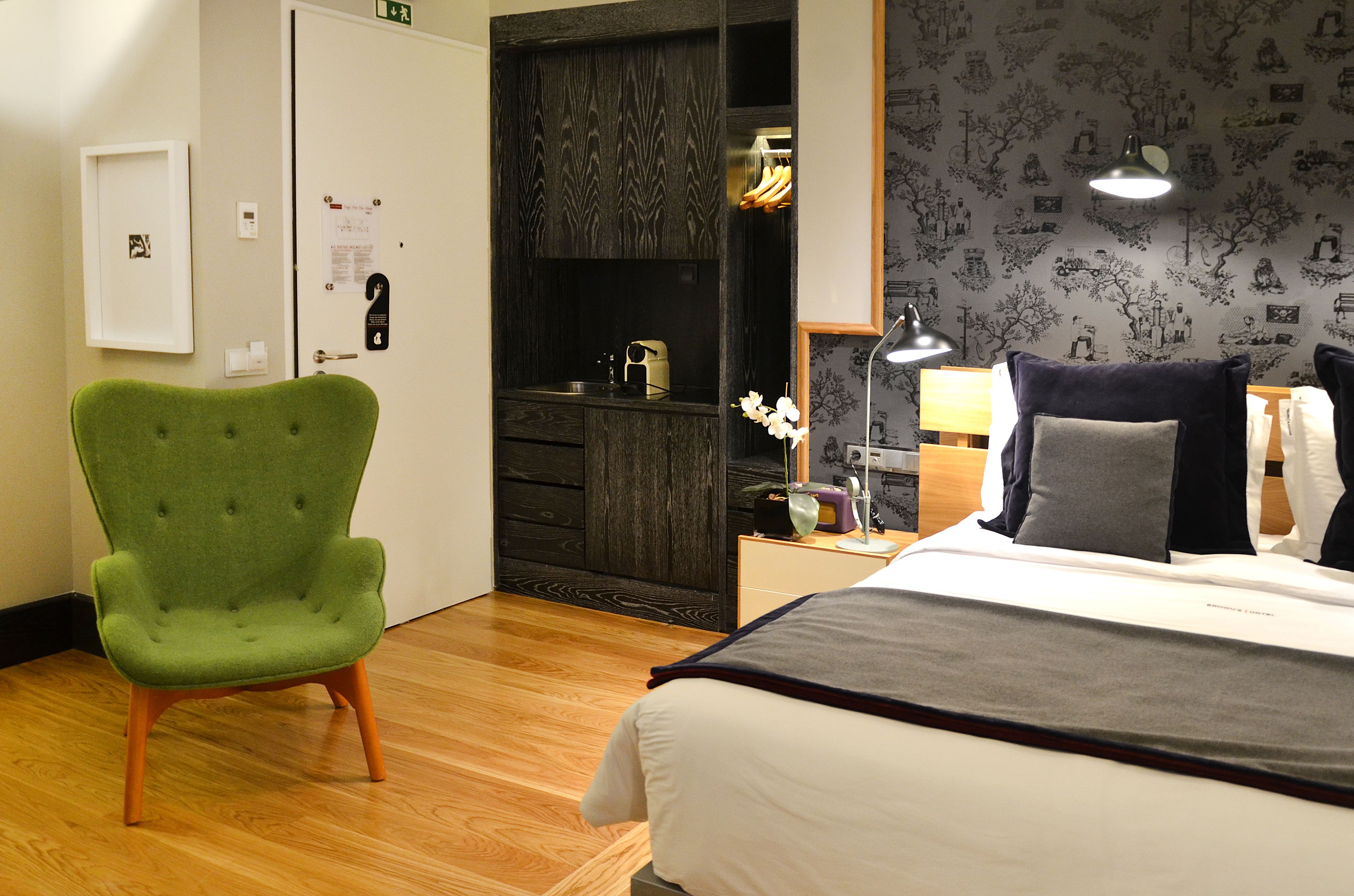 Une chambre d’hôtel avec un lit, un fauteuil vert, une commode en bois, une table de chevet avec une lampe et un mur orné d’un motif floral.