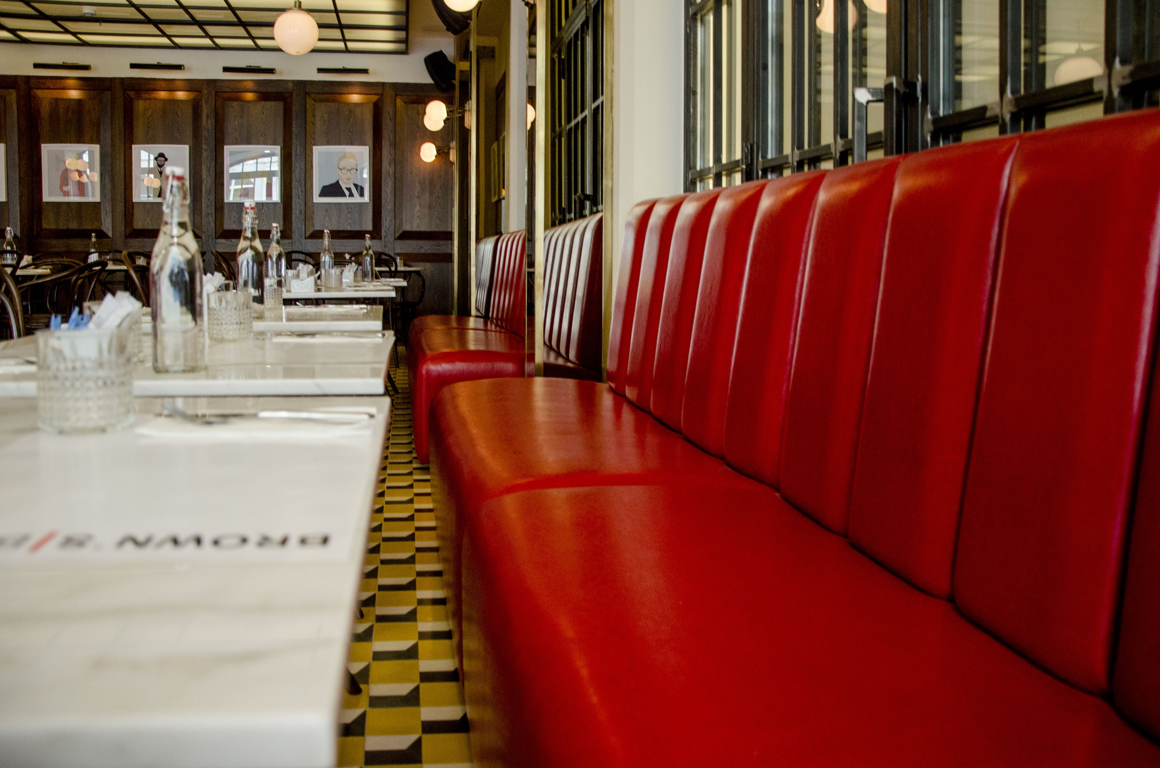 Interior de restaurante con asientos tapizados en rojo, piso a cuadros y mesas dispuestas con botellas y vasos.