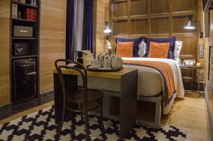 Et hotelværelse med trægulv, en seng med orange og blå puder, et skrivebord med stol, en reol og et lille bord med lampe.