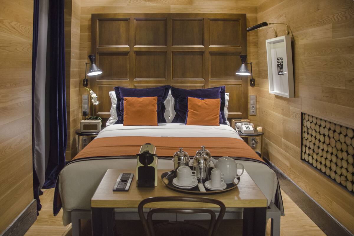 Habitación de hotel con cabecera de madera, cama con ropa de cama naranja y blanca, mesita de noche con teléfono y bandeja con alimentos para el desayuno.