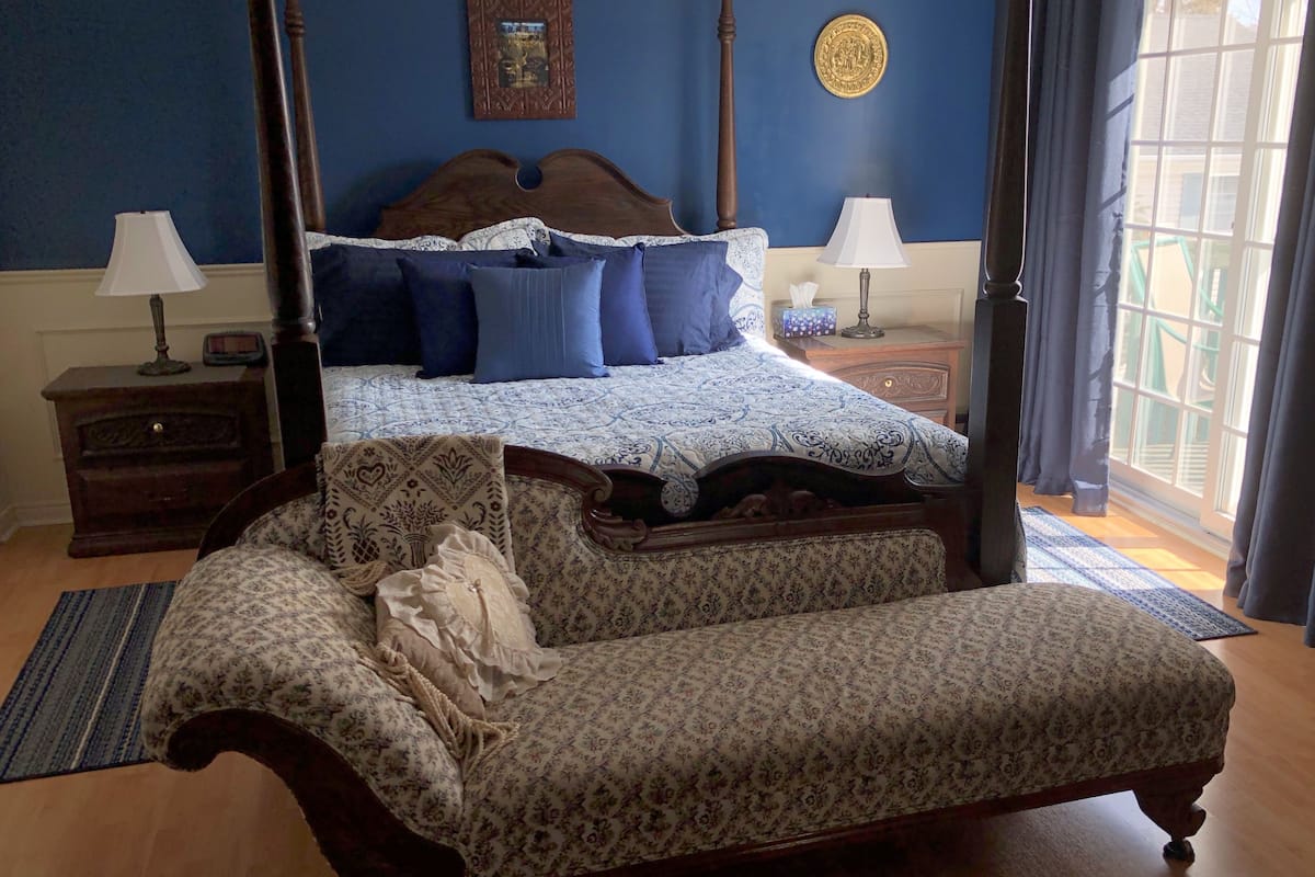 A four-poster bed with a blue and white patterned bedspread, a wooden nightstand with a lamp, and a large window with curtains.