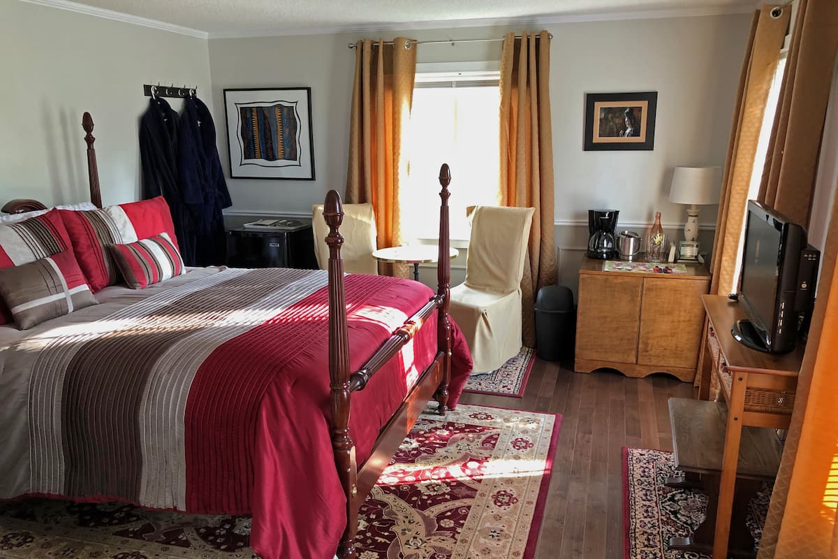 A neatly arranged bedroom with a four-poster bed, a chair, a desk with a lamp, and a television.