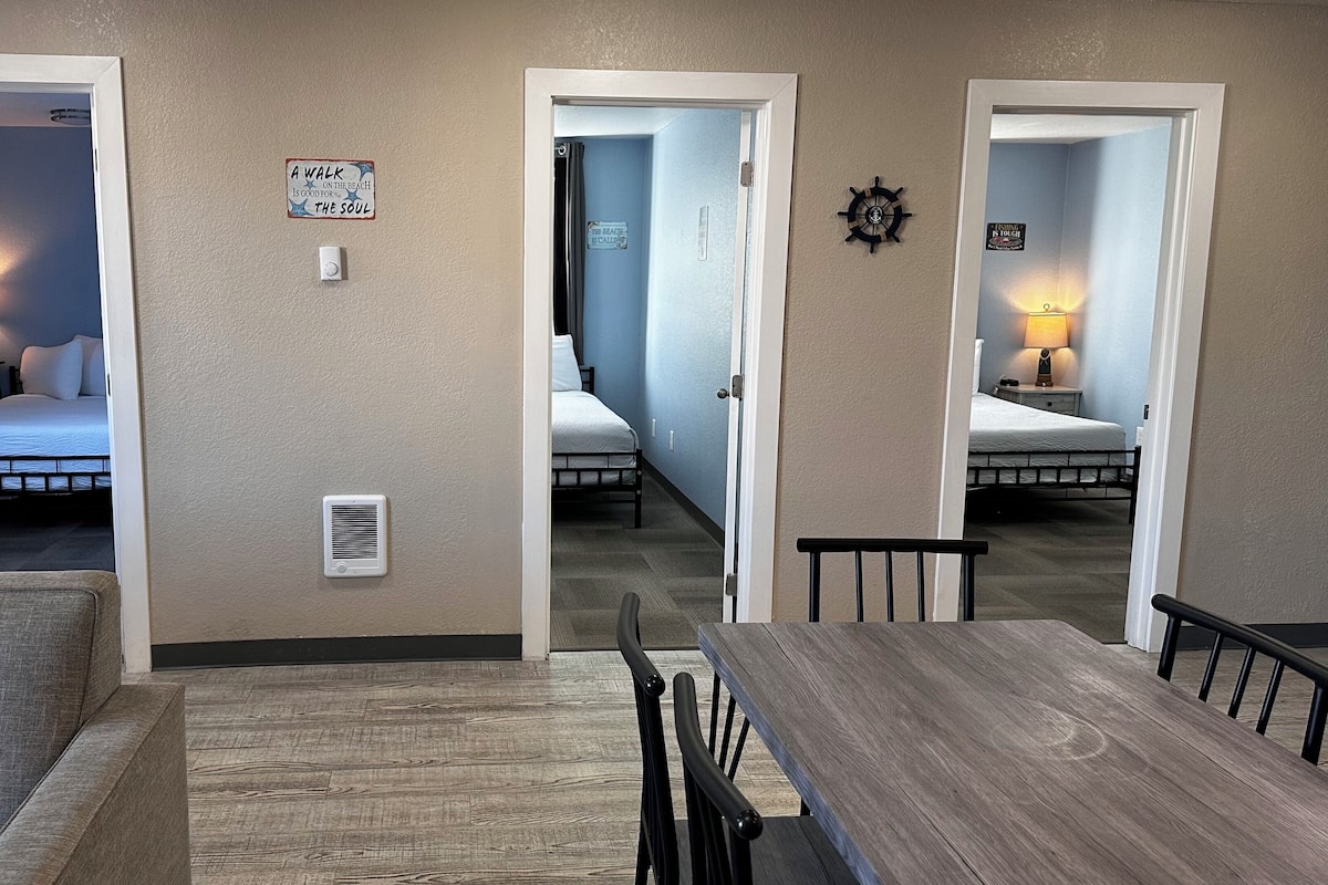 Habitación de hotel con una mesa de comedor de madera y sillas, una cama con colcha blanca y una mesita de noche con lámpara.