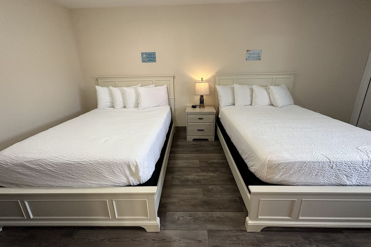 Una habitación con dos camas, cada una con ropa blanca y cabeceros, una mesita de noche con lámpara y una pequeña señal en la pared.