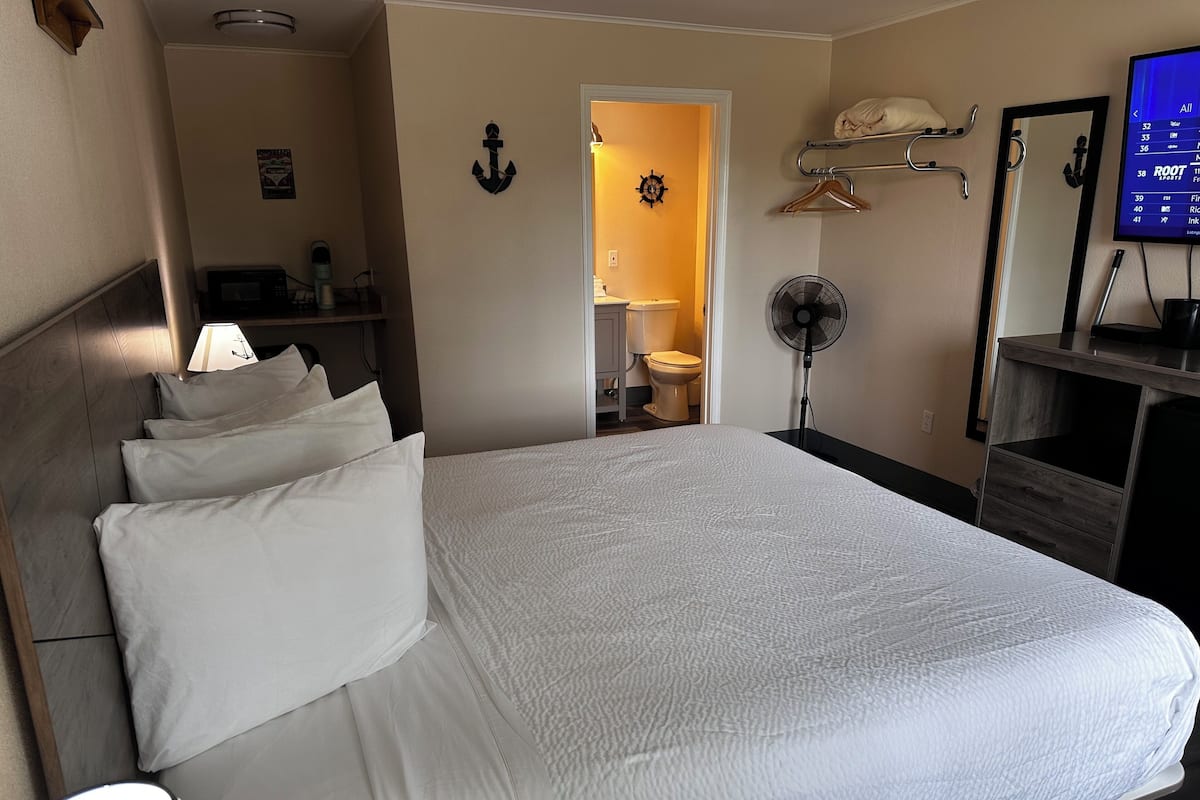 Habitación de hotel con una cama grande, una mesita de noche con lámpara, un ventilador, un espejo y un televisor en la pared.