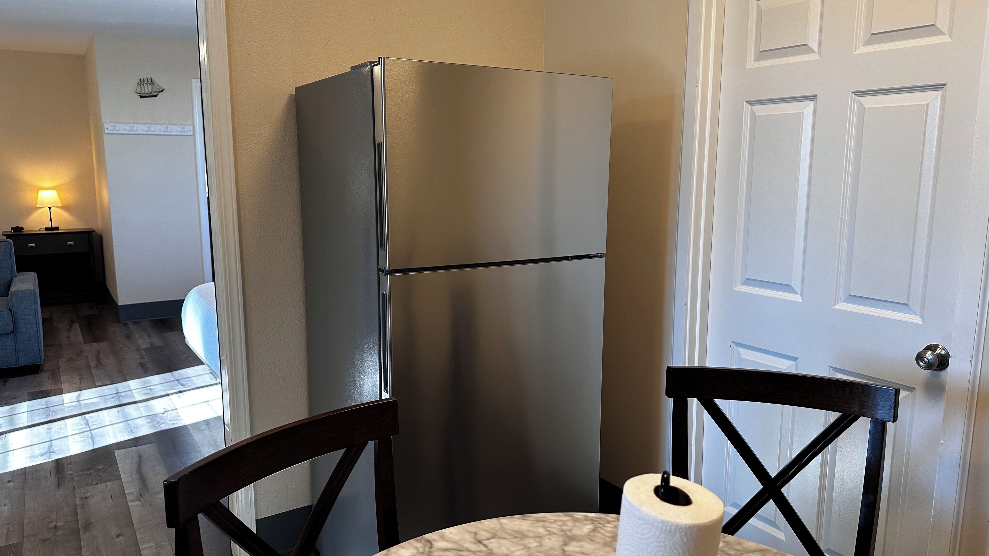 A compact kitchen with a stainless steel refrigerator, a wooden dining table with chairs, and a white door in the background.