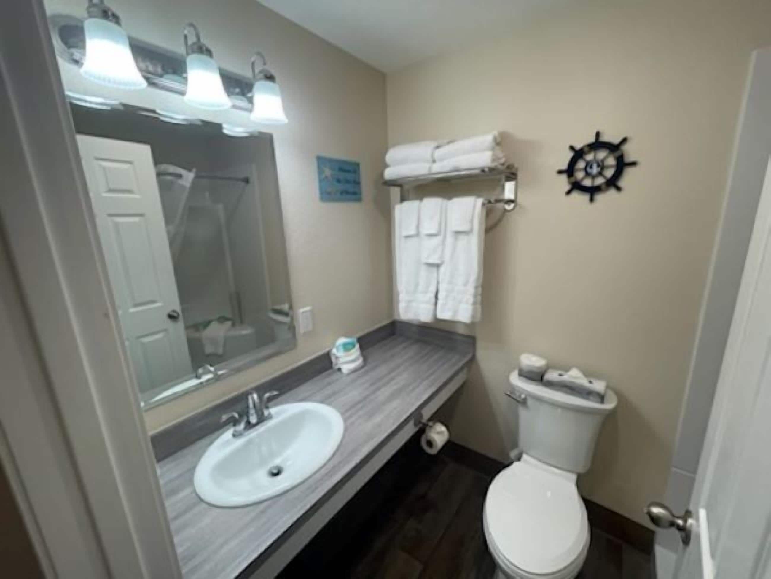 A bathroom with a white sink, a toilet, and a large mirror. There are towels hanging on the wall and a ship's wheel decoration.